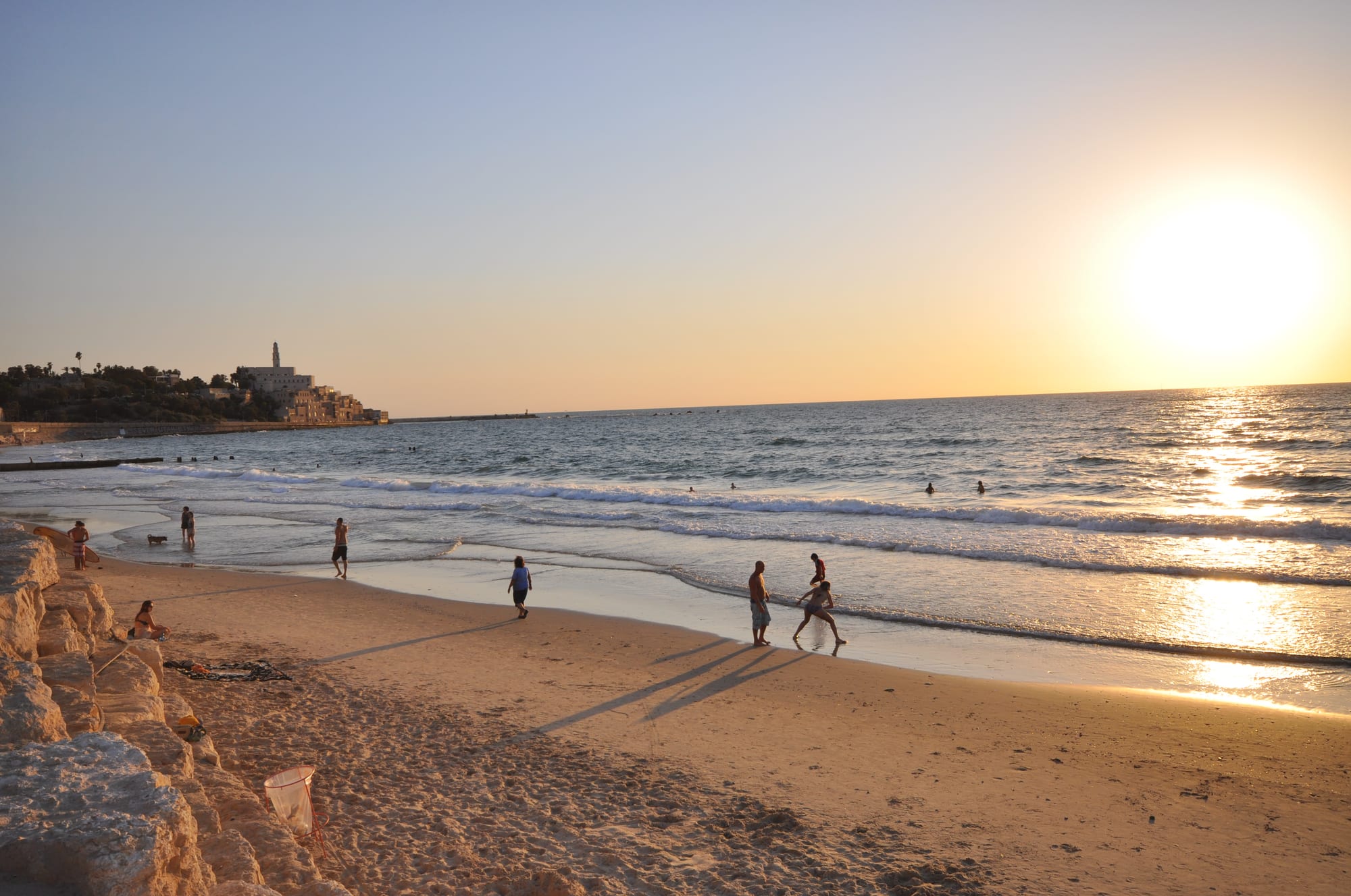 People walking and swimming at a Tel Aviv beach at sunset, with the old city of Jaffa visible in the distance