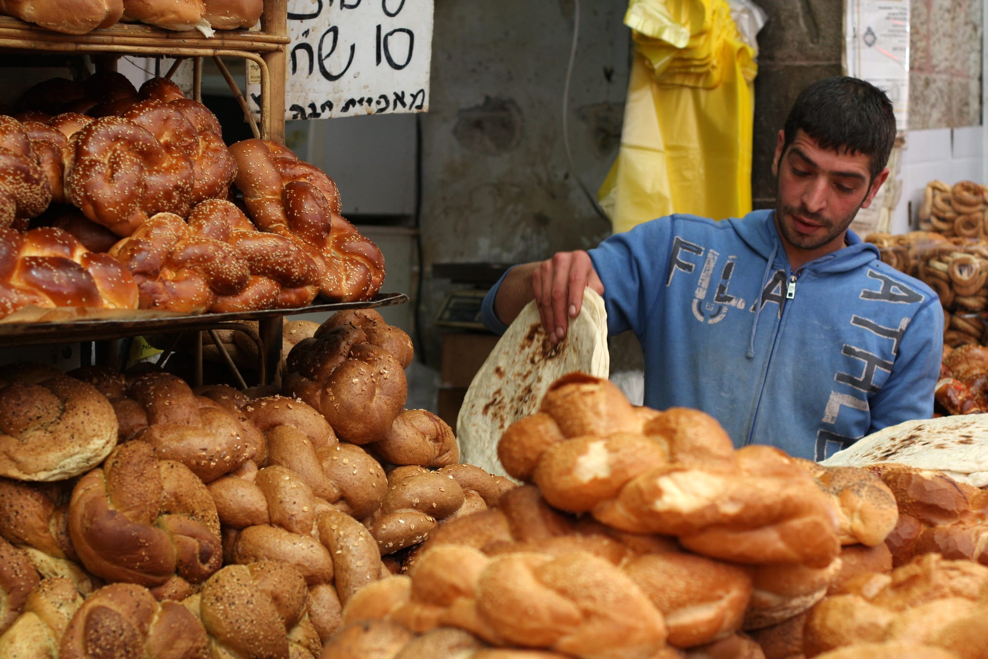 Stacks of fresh challah and other bread loaves piled on a market stall, with a vendor arranging them