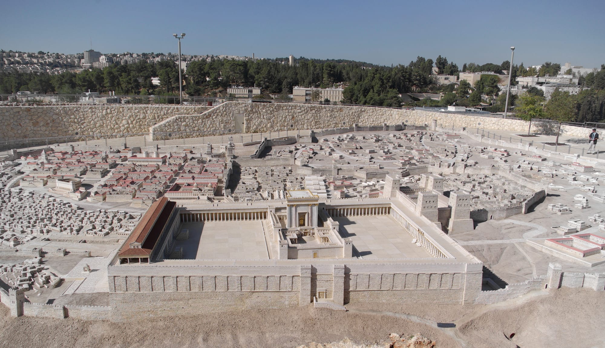 A large outdoor scale model of an ancient walled city in pale stone, with a colonnaded temple compound in the foreground and modern buildings and trees beyond