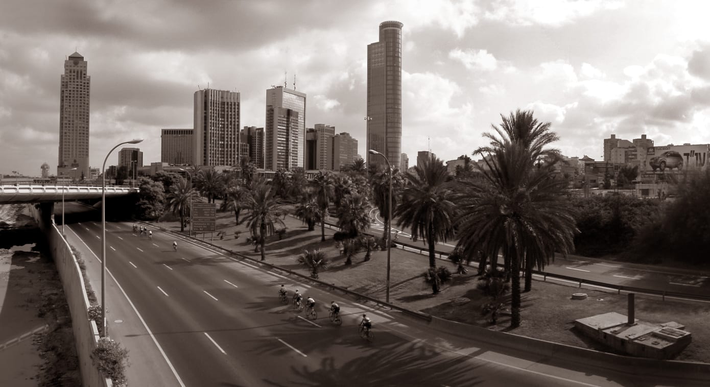 A sepia-toned view of a wide urban highway empty of cars except for a small group of cyclists, with palm trees and office towers alongside