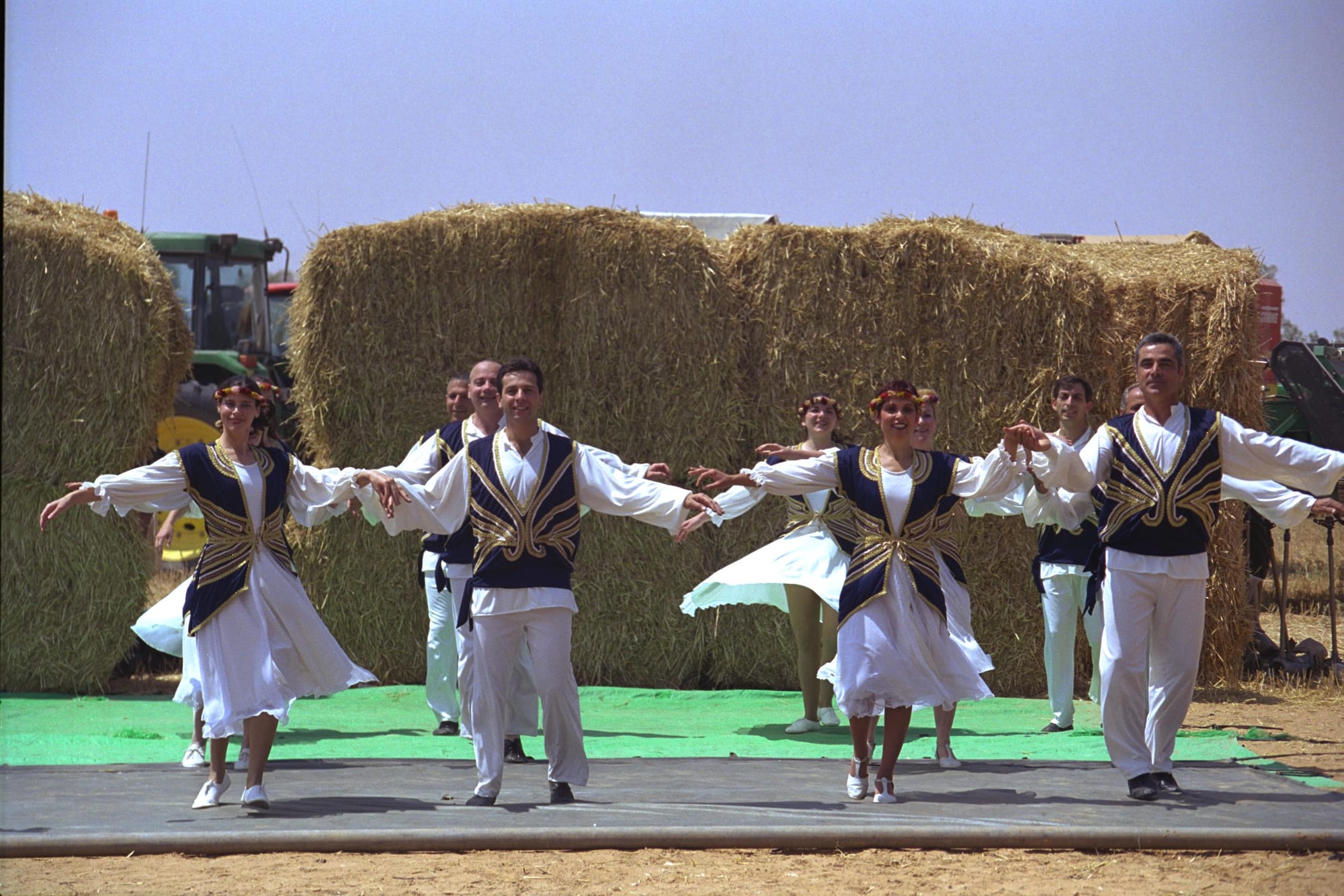 Dancers in white costumes with dark embroidered waistcoats and flower garlands perform with arms outstretched on an outdoor stage in front of large hay bales and a tractor