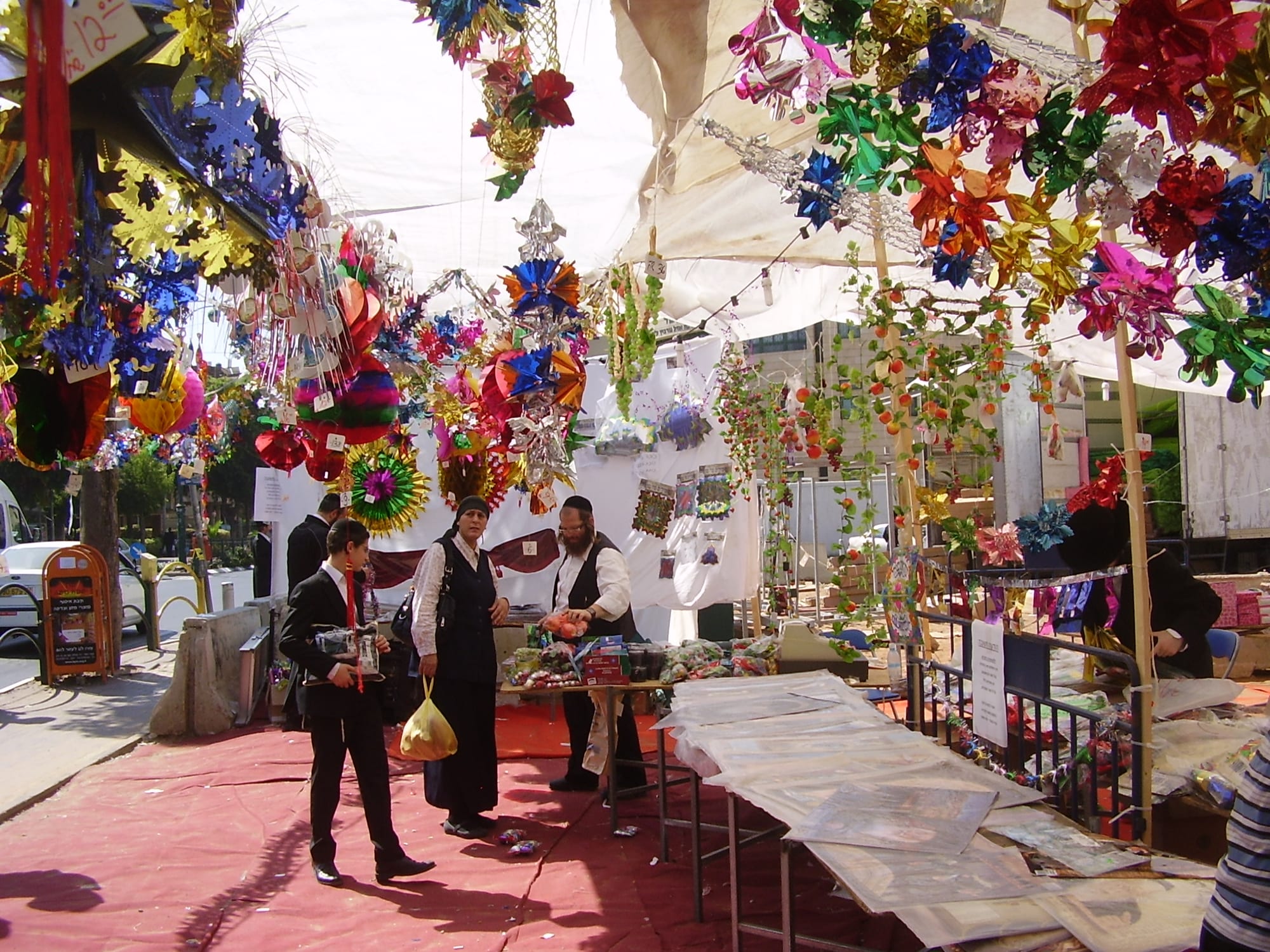 A street market under awnings hung with brightly coloured paper and foil decorations, with shoppers in dark clothing at a stall