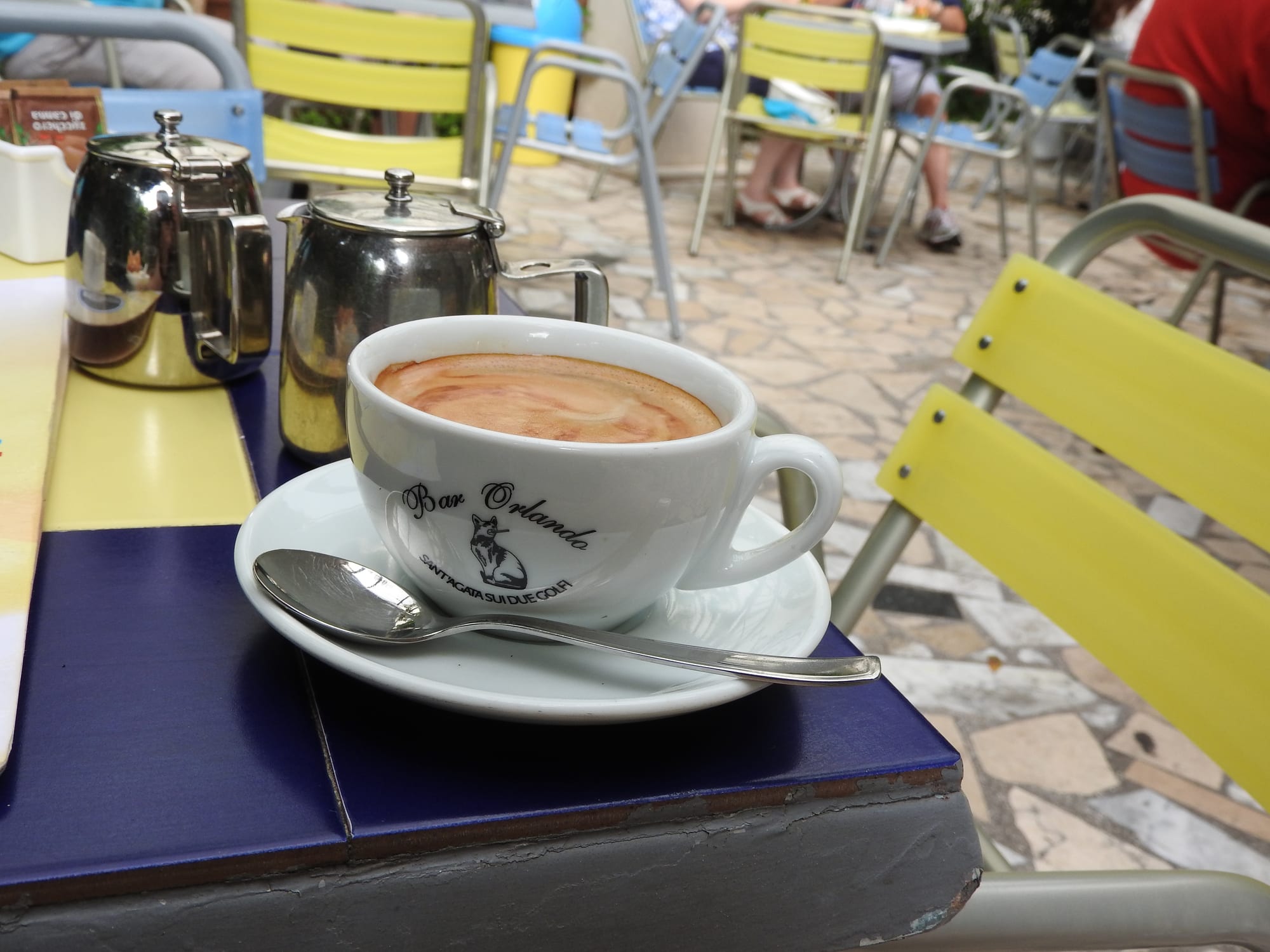 A cappuccino served at an outdoor café table with yellow metal chairs, the cup marked Bar Orlando