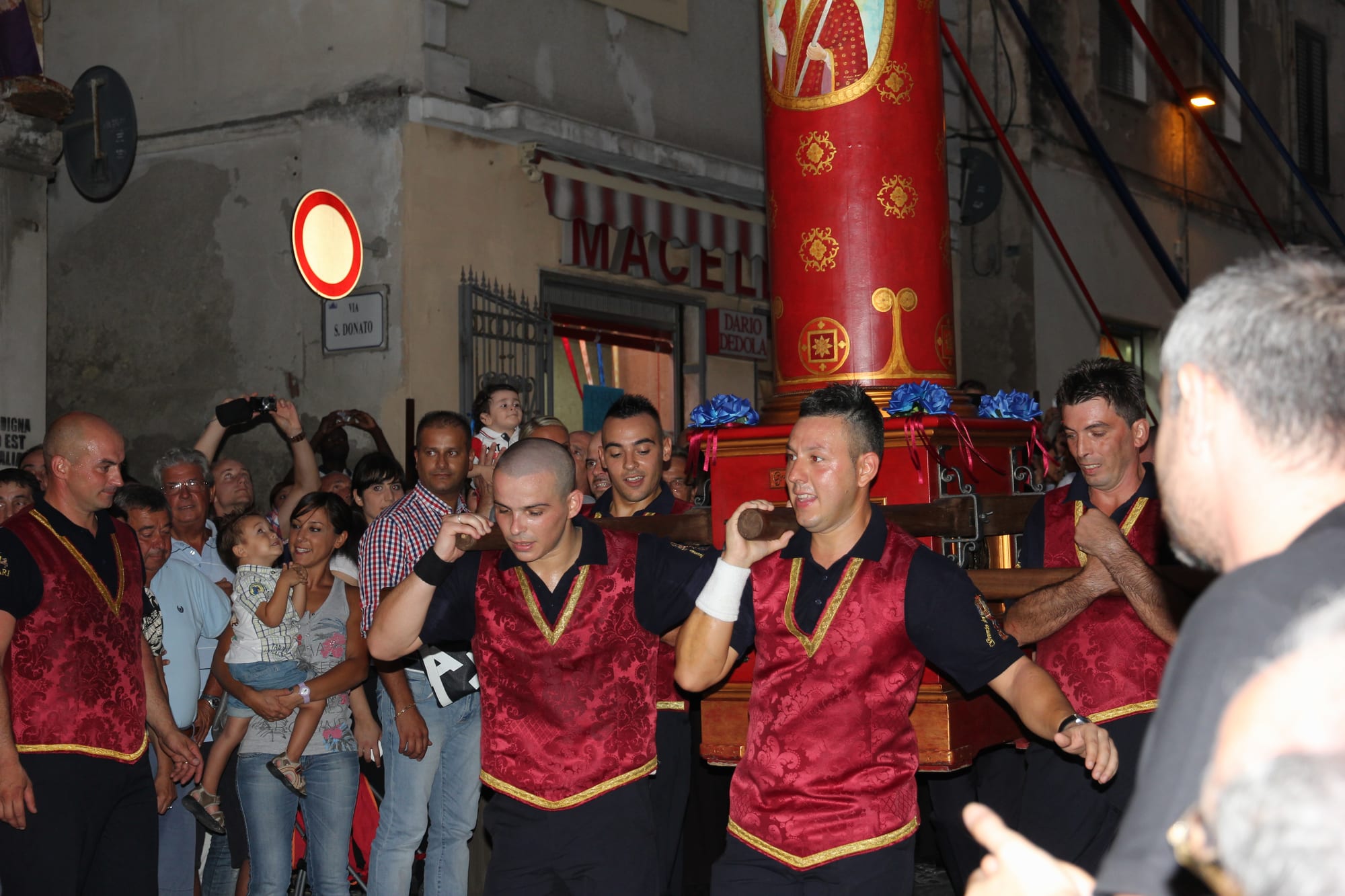 Men in matching red brocade waistcoats carry a tall red and gold decorated column on wooden poles through a narrow street at dusk, watched by a crowd