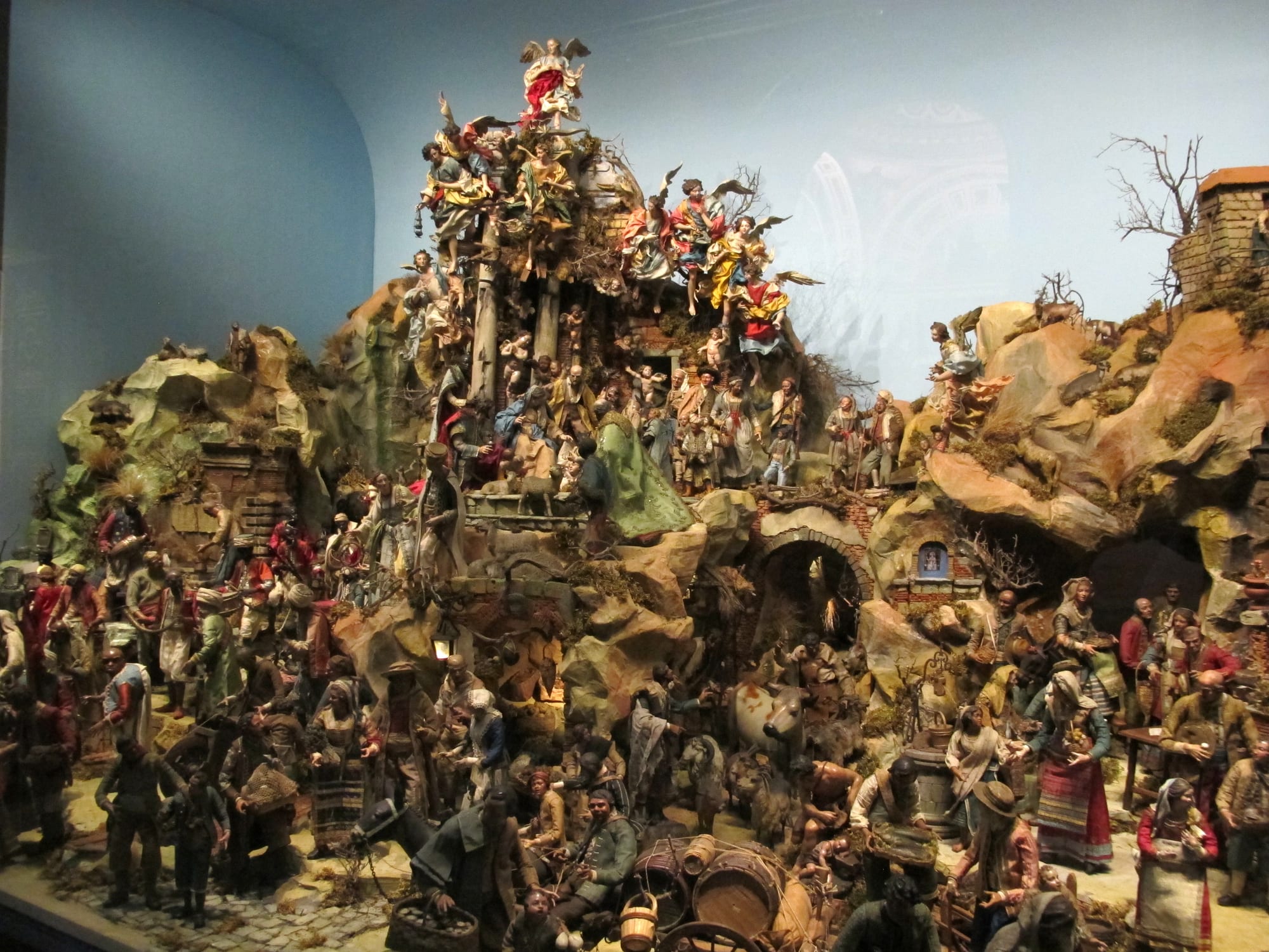 A large and crowded nativity scene of hand-painted figures on rocky tiers, with angels in bright robes at the top and villagers, animals and market stalls below