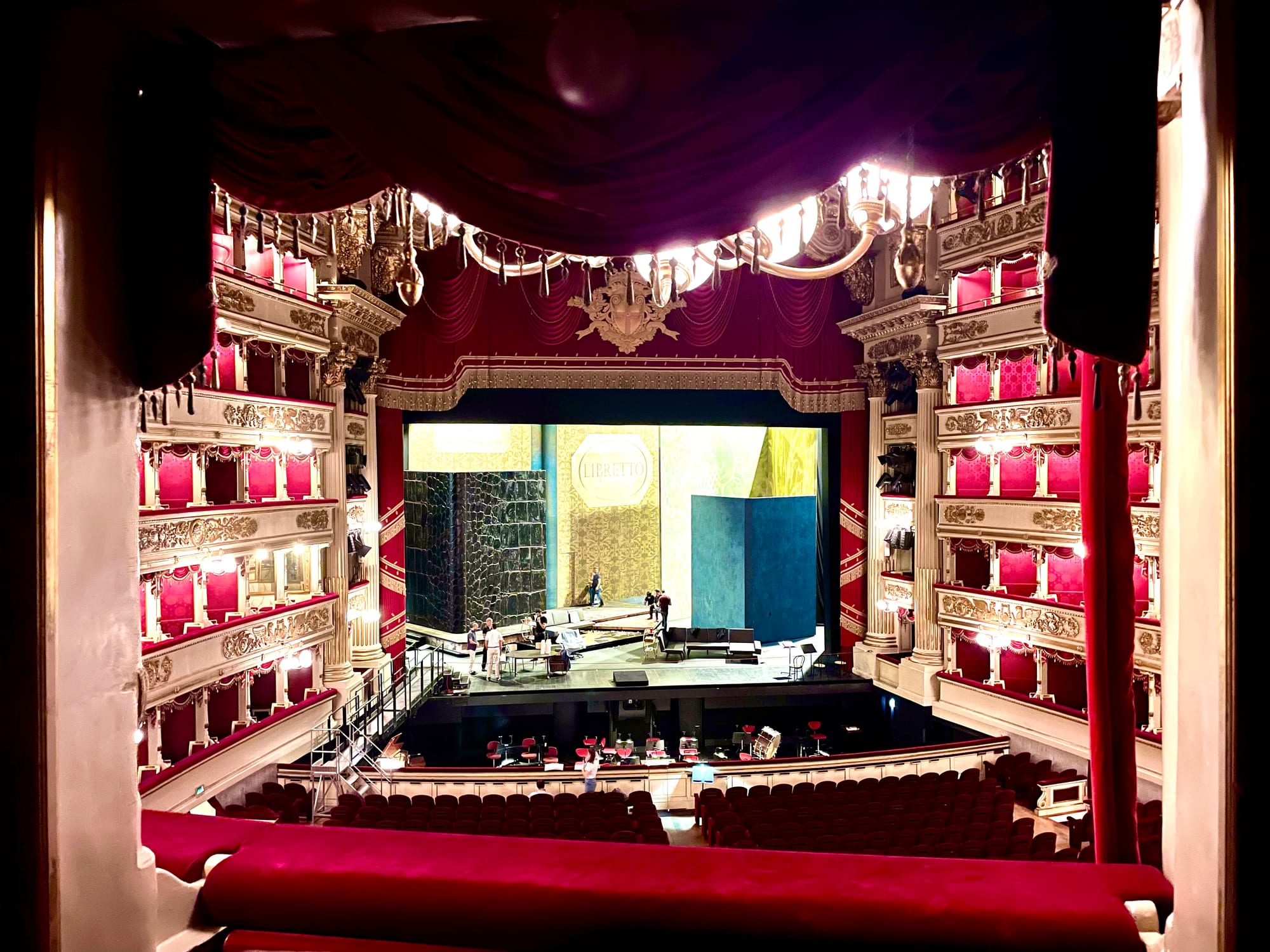 A red and gold opera house auditorium seen from a box, with tiers of boxes curving around a lit stage where a set is being prepared