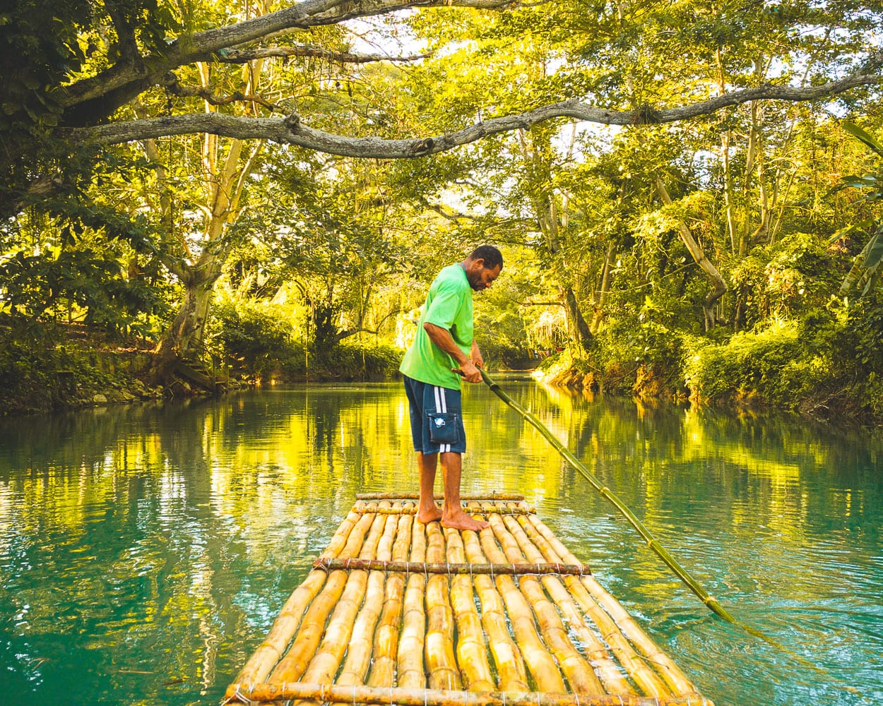 A bamboo raft with two passengers being poled down a calm jungle-lined river