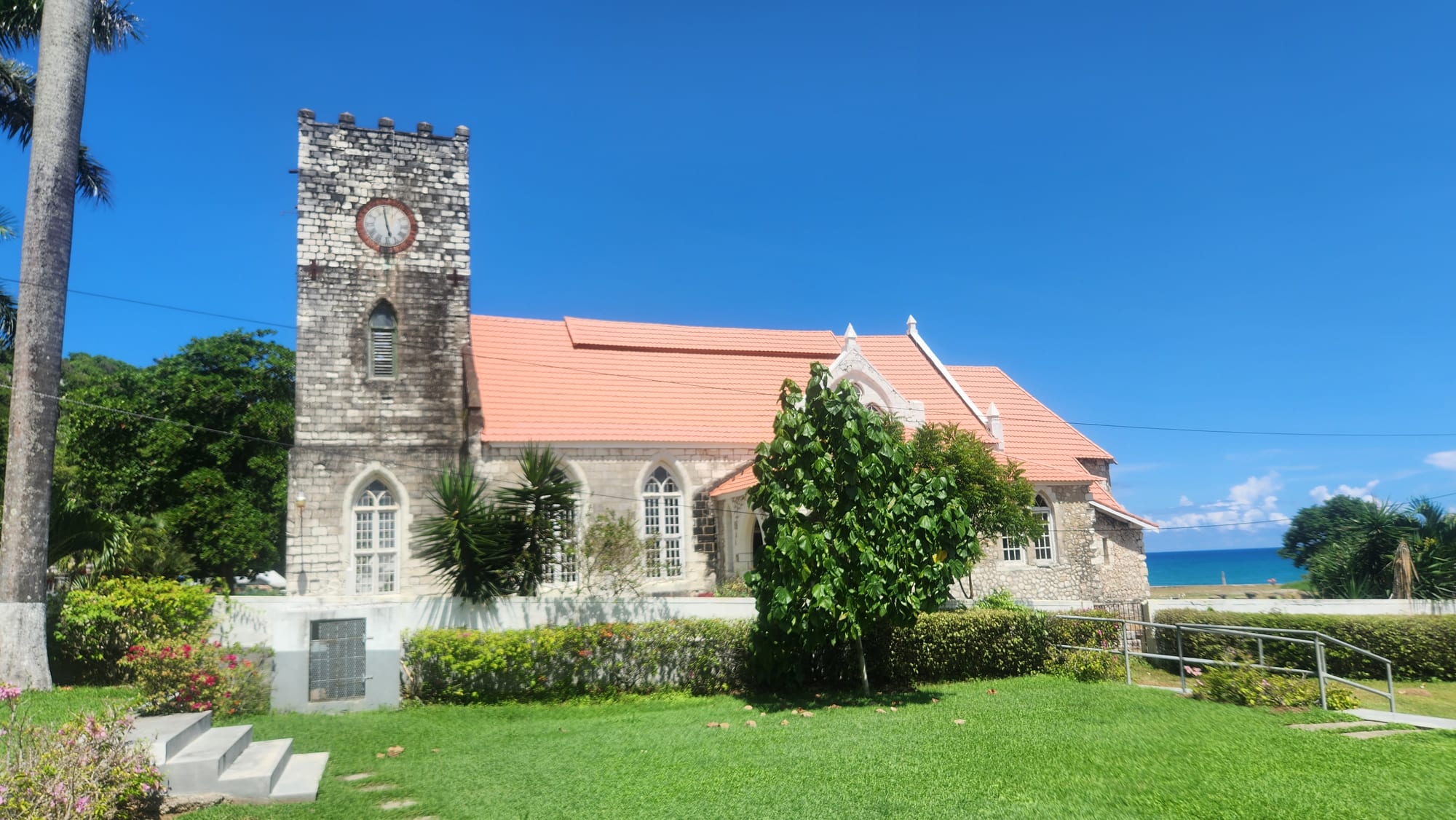 A stone church with a square clock tower and a salmon-pink tiled roof on a green lawn with palm trees, the sea visible beyond
