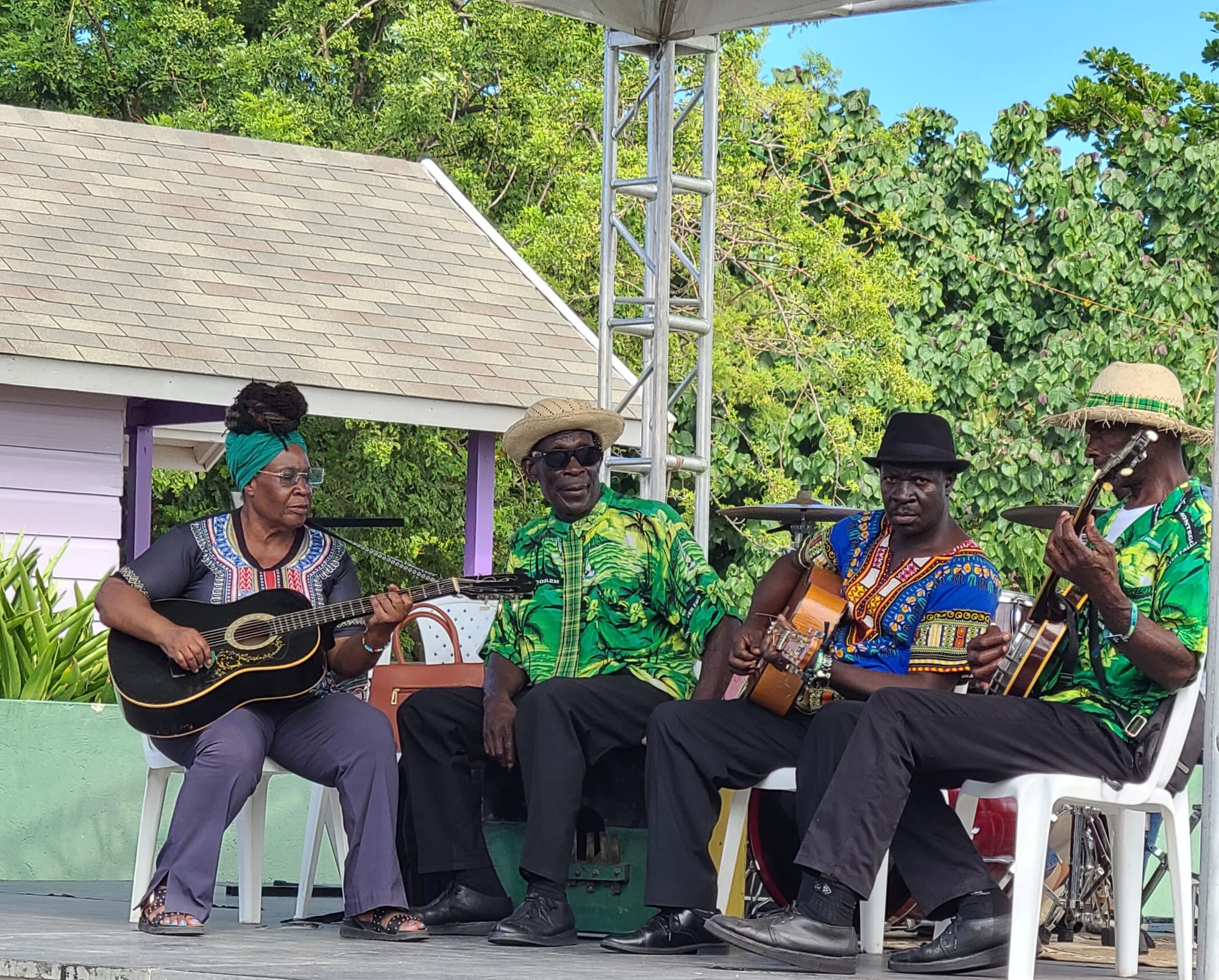 Four musicians seated on an outdoor stage playing acoustic guitars and a banjo, most wearing hats and brightly patterned shirts, with trees behind