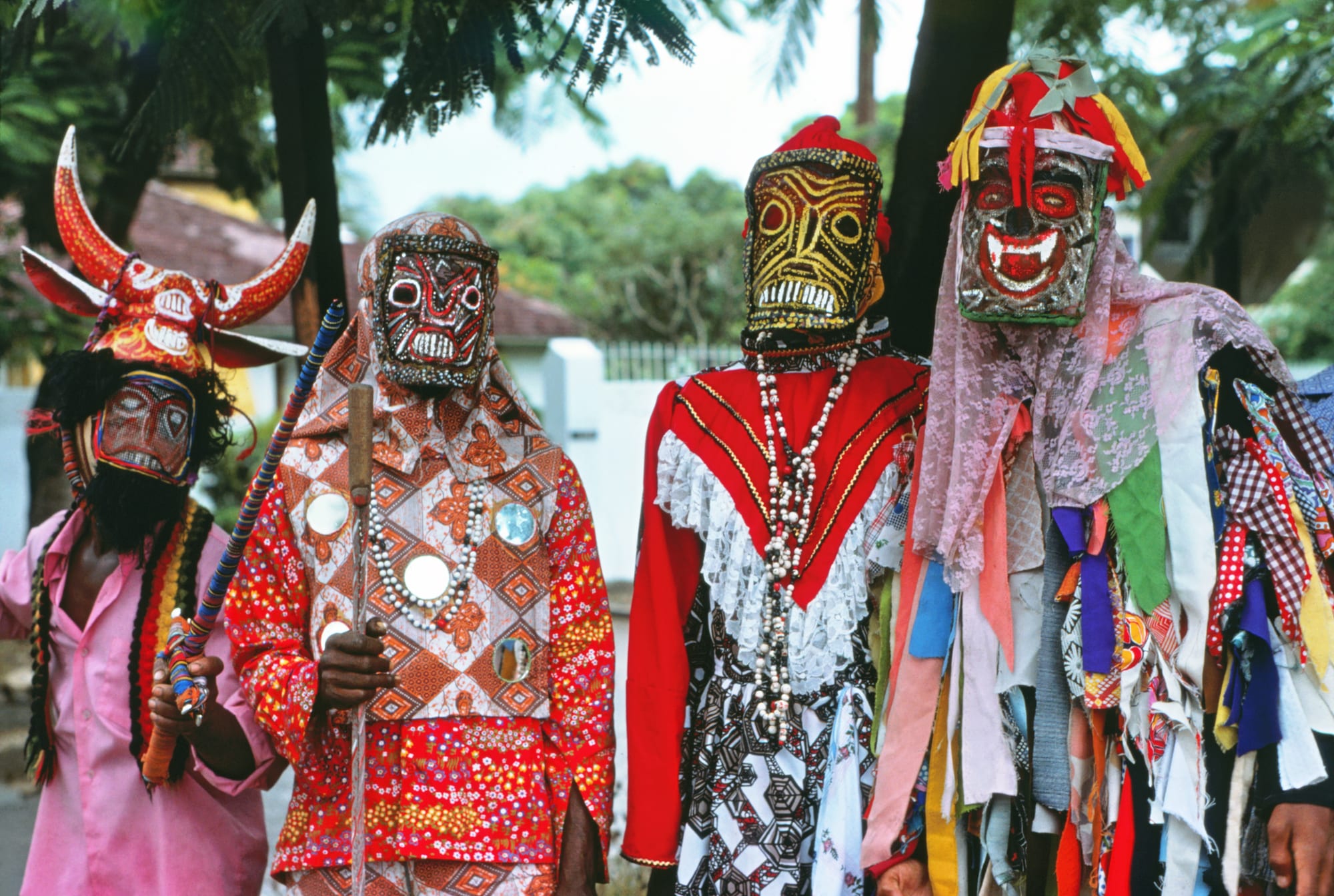 Four masked masqueraders in brightly patterned costumes of cloth, lace and ribbons, one with a horned red headdress, standing together under trees