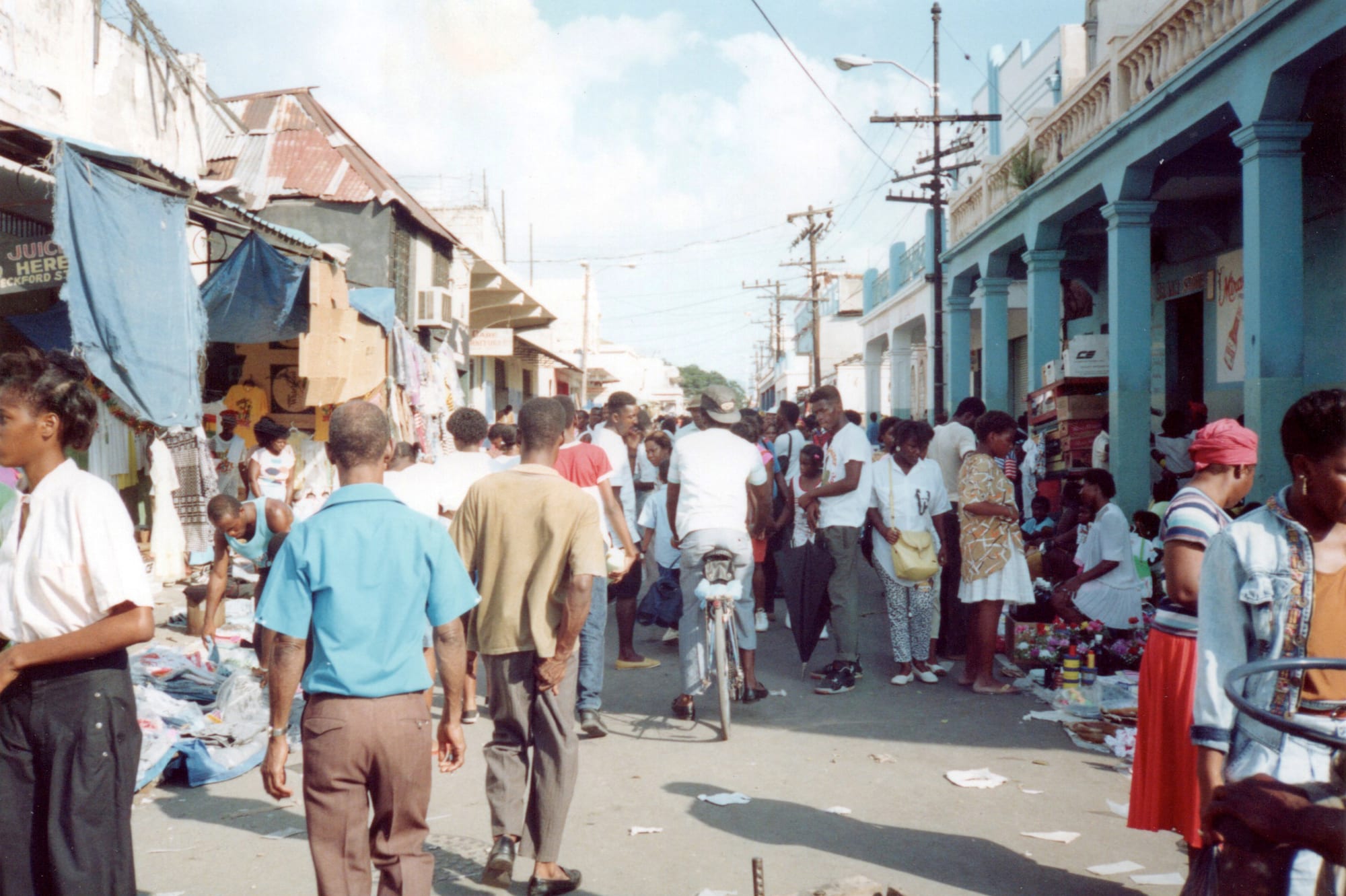 A crowded downtown Kingston street lined with vendors and shoppers, goods laid out on the pavement outside colonnaded shop buildings