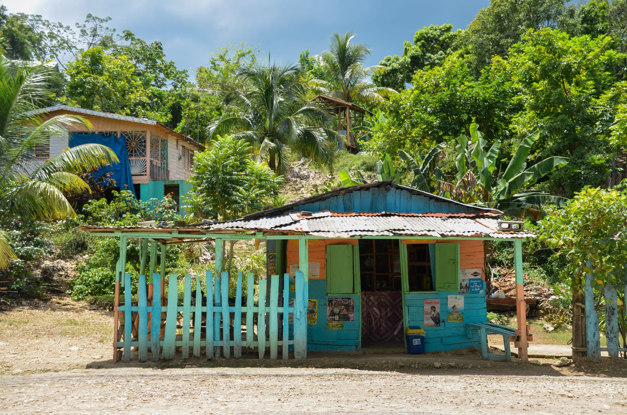 A small blue and orange wooden roadside bar with a house visible behind it among palm trees