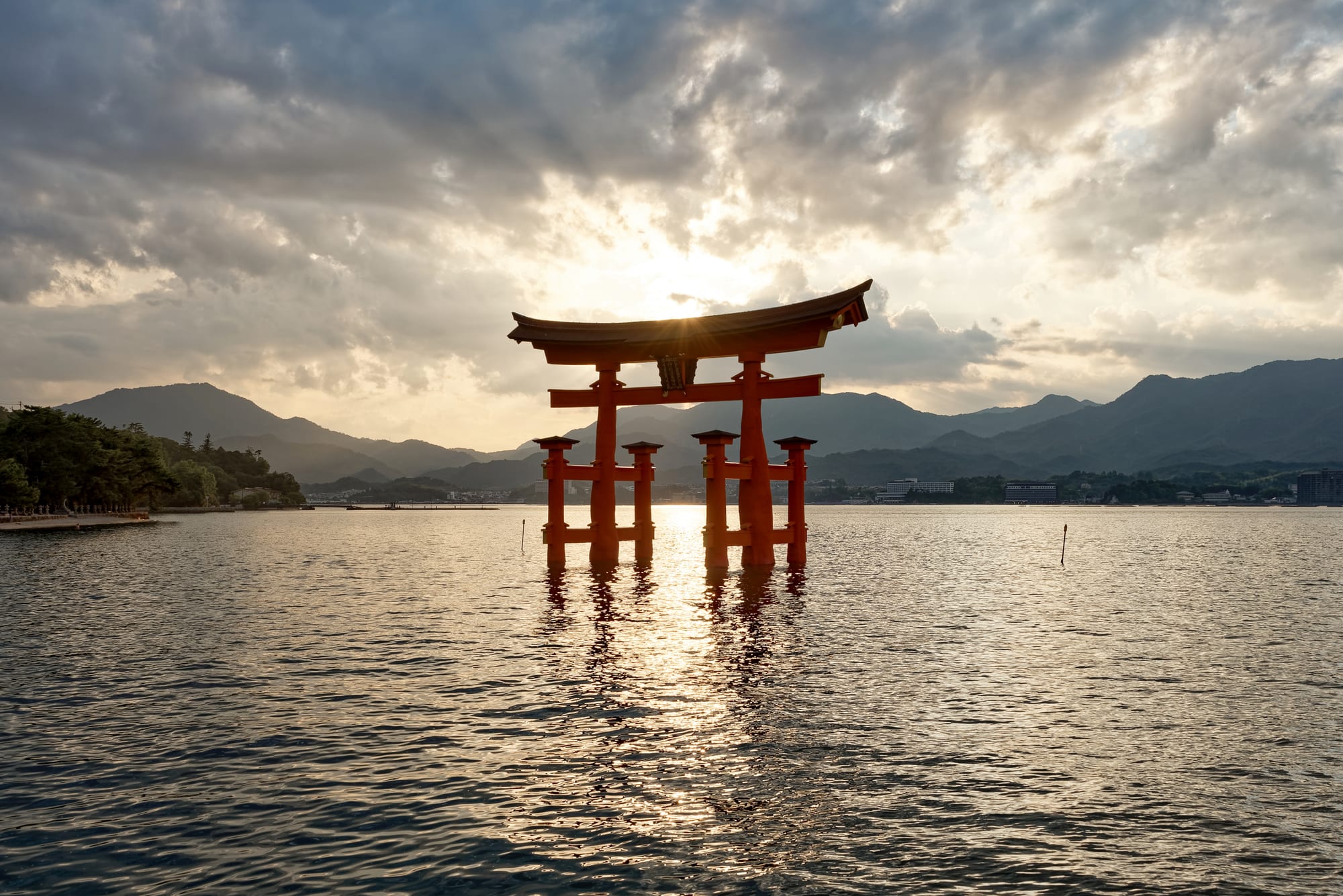 A large vermilion torii gate standing in the sea at sunset, a shrine on the shore behind