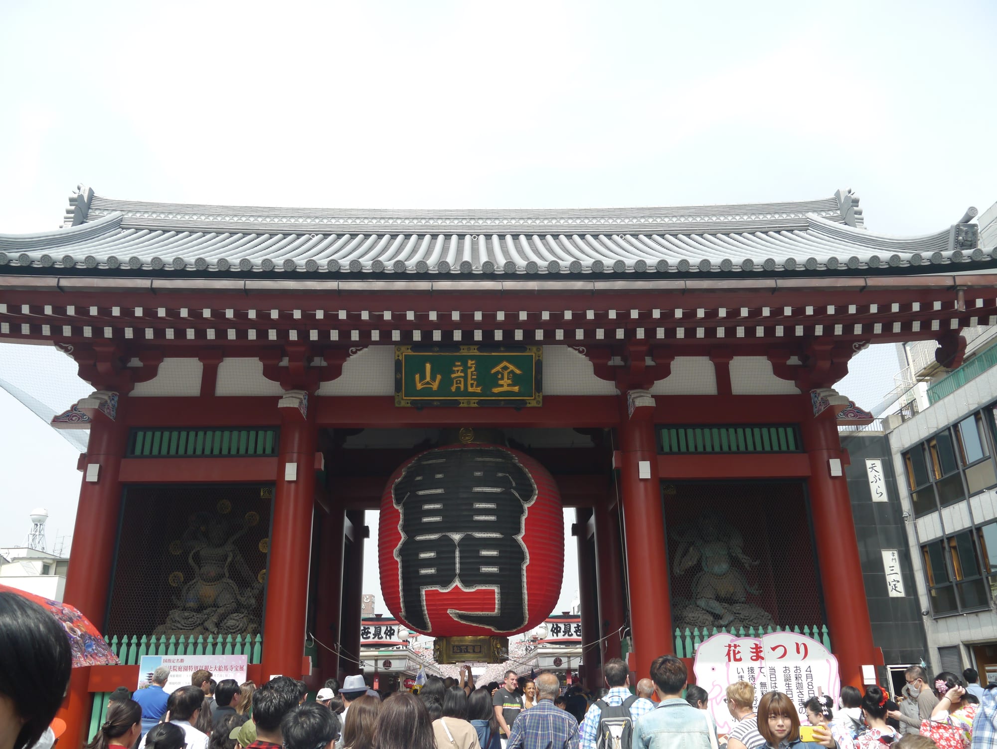 A large red temple gate with a giant paper lantern, crowds passing beneath