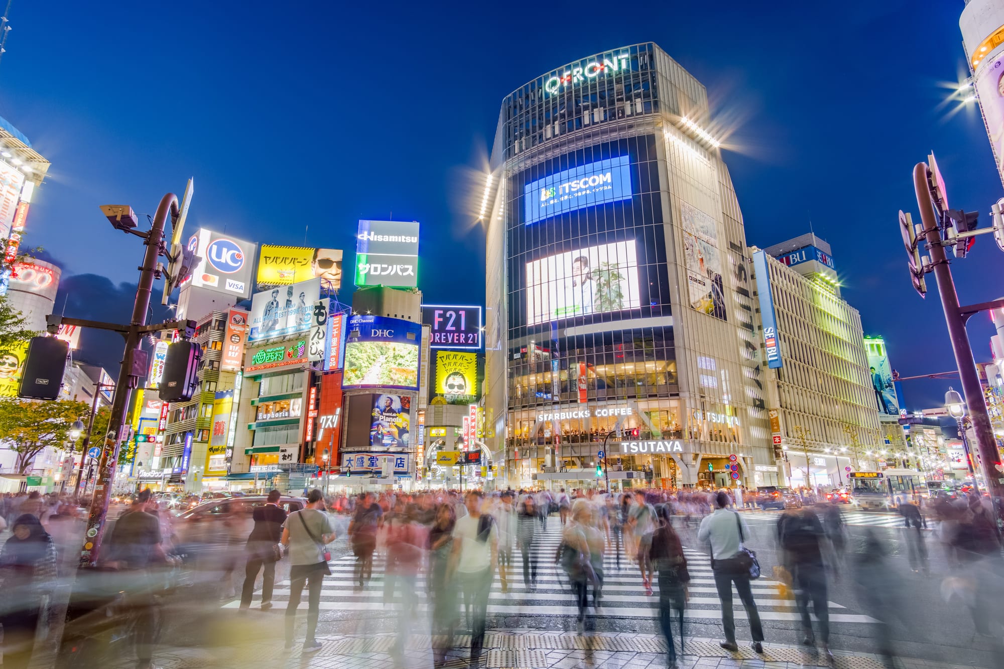 A massive pedestrian intersection packed with crowds crossing in all directions, ringed by video screens