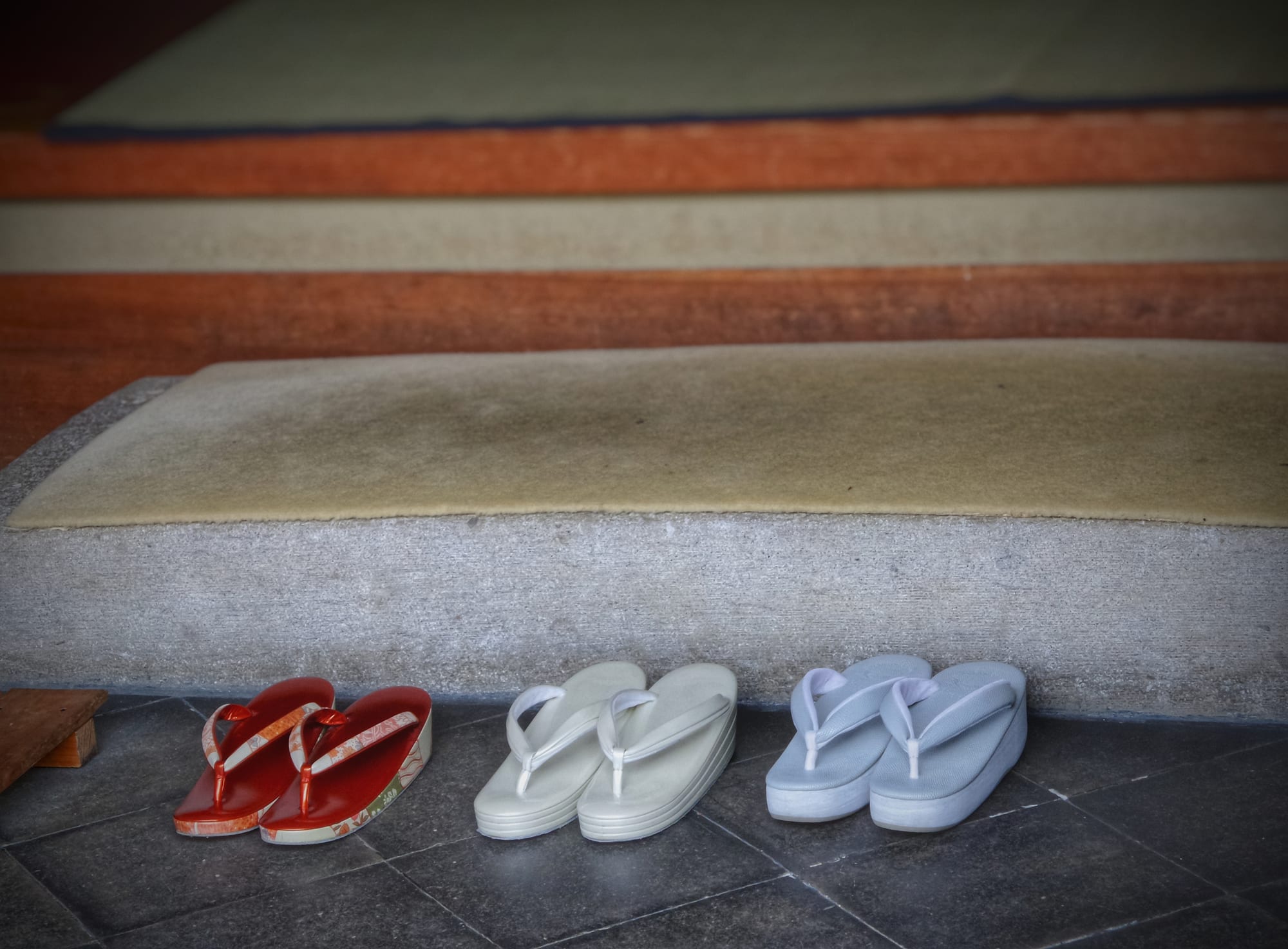 Three pairs of traditional thonged sandals, one red and two pale, lined up neatly on a stone floor below a raised step leading to tatami mats