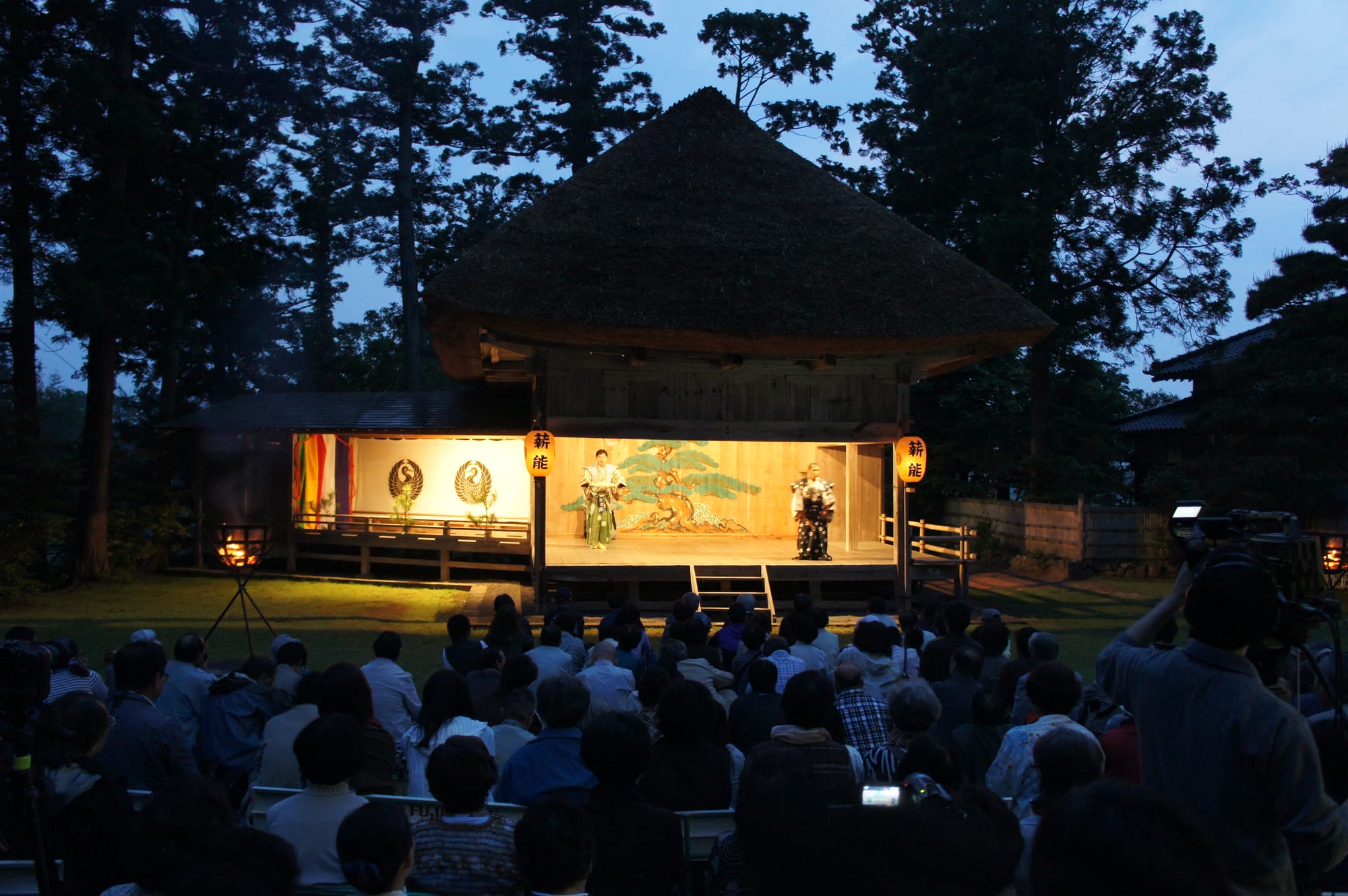 Two costumed performers on a lit wooden stage under a thatched roof at dusk, with a painted pine tree on the back wall, lanterns on either side, fire baskets and a seated audience in the foreground