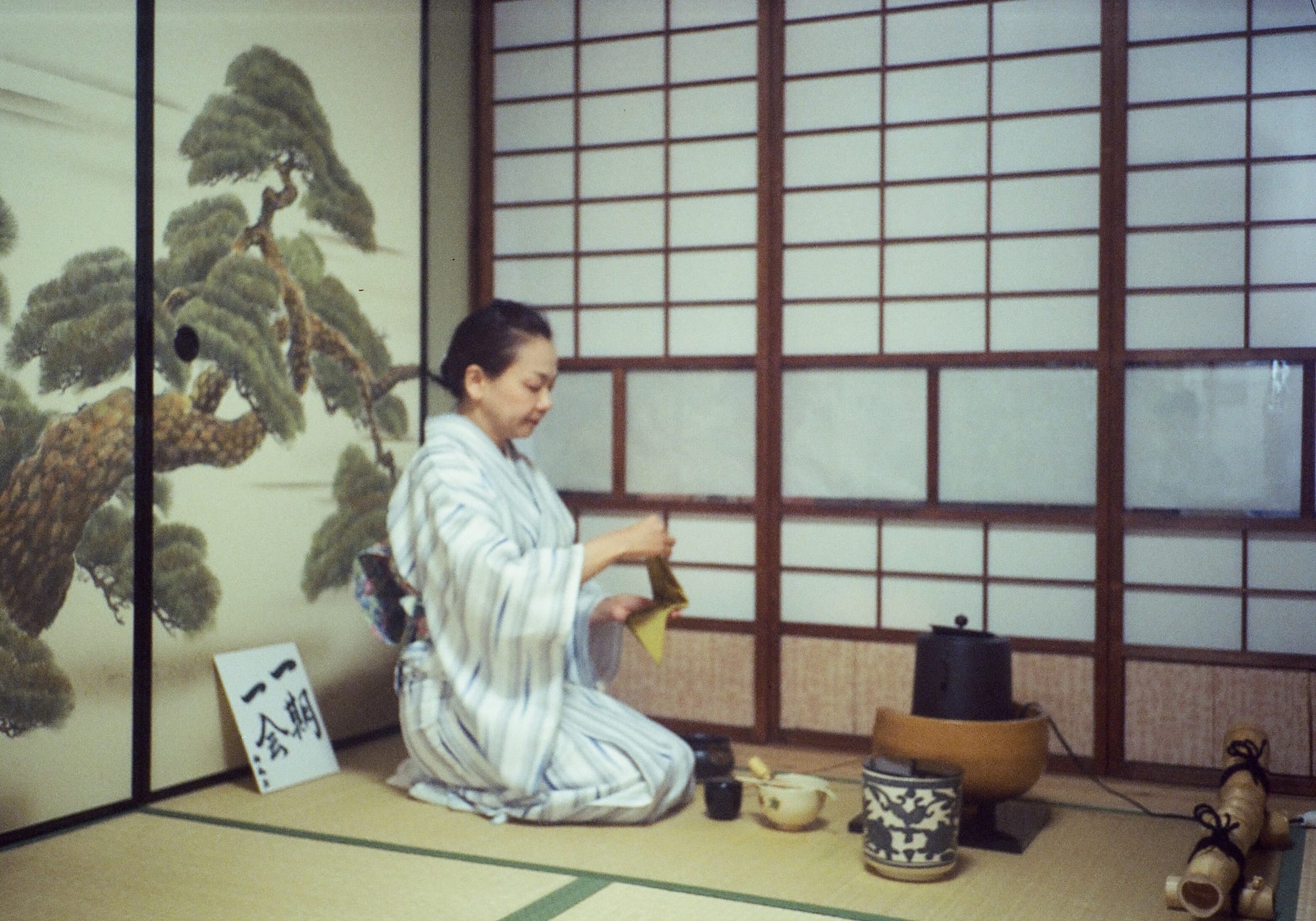 A woman in a striped kimono kneels on tatami mats in front of paper shoji screens, holding a small folded cloth, with an iron kettle, a blue and white water jar and a tea bowl with a bamboo whisk set out beside her