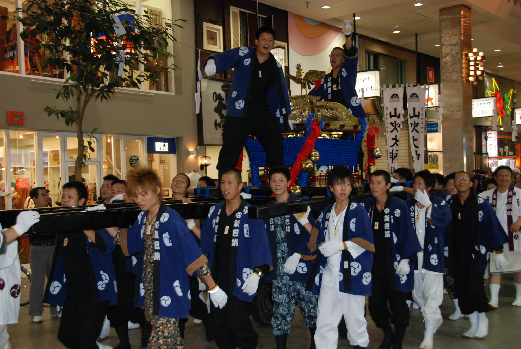 A team of young men in matching blue and white happi coats and white gloves shouldering the black poles of a gilded portable shrine through a lit shopping arcade, with two men standing on top of it