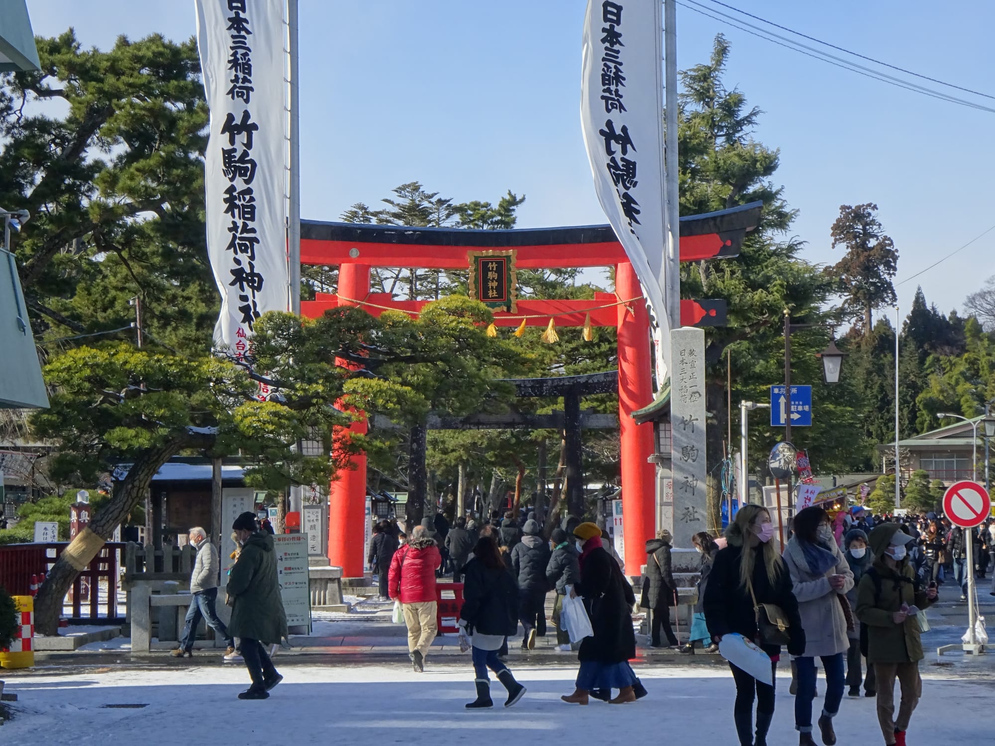 A large red torii gate framed by pine trees and tall white banners with black lettering, with people in winter coats walking along the approach towards and away from it