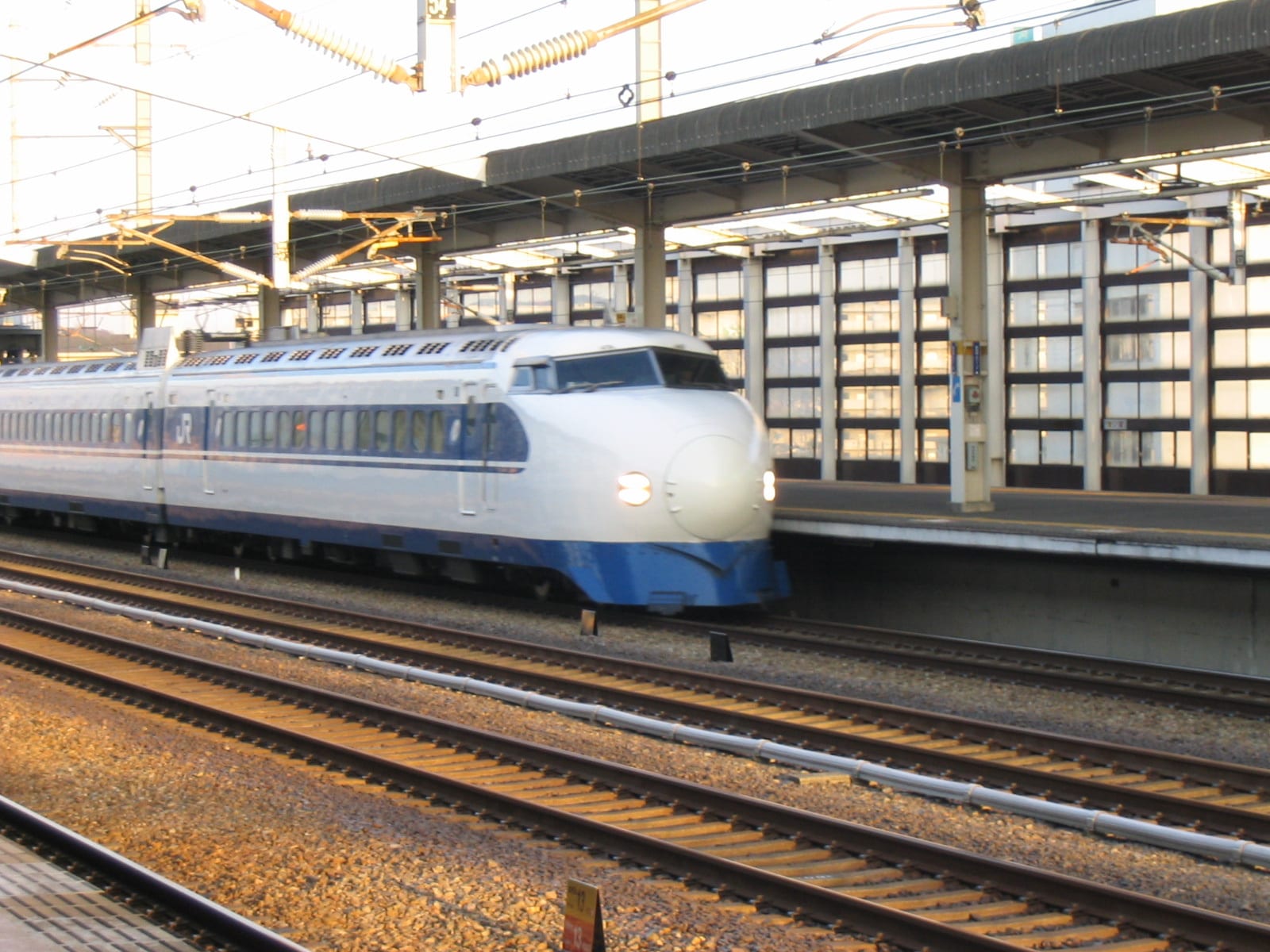 A white and blue bullet train with a rounded nose pulling through a station beside several railway tracks