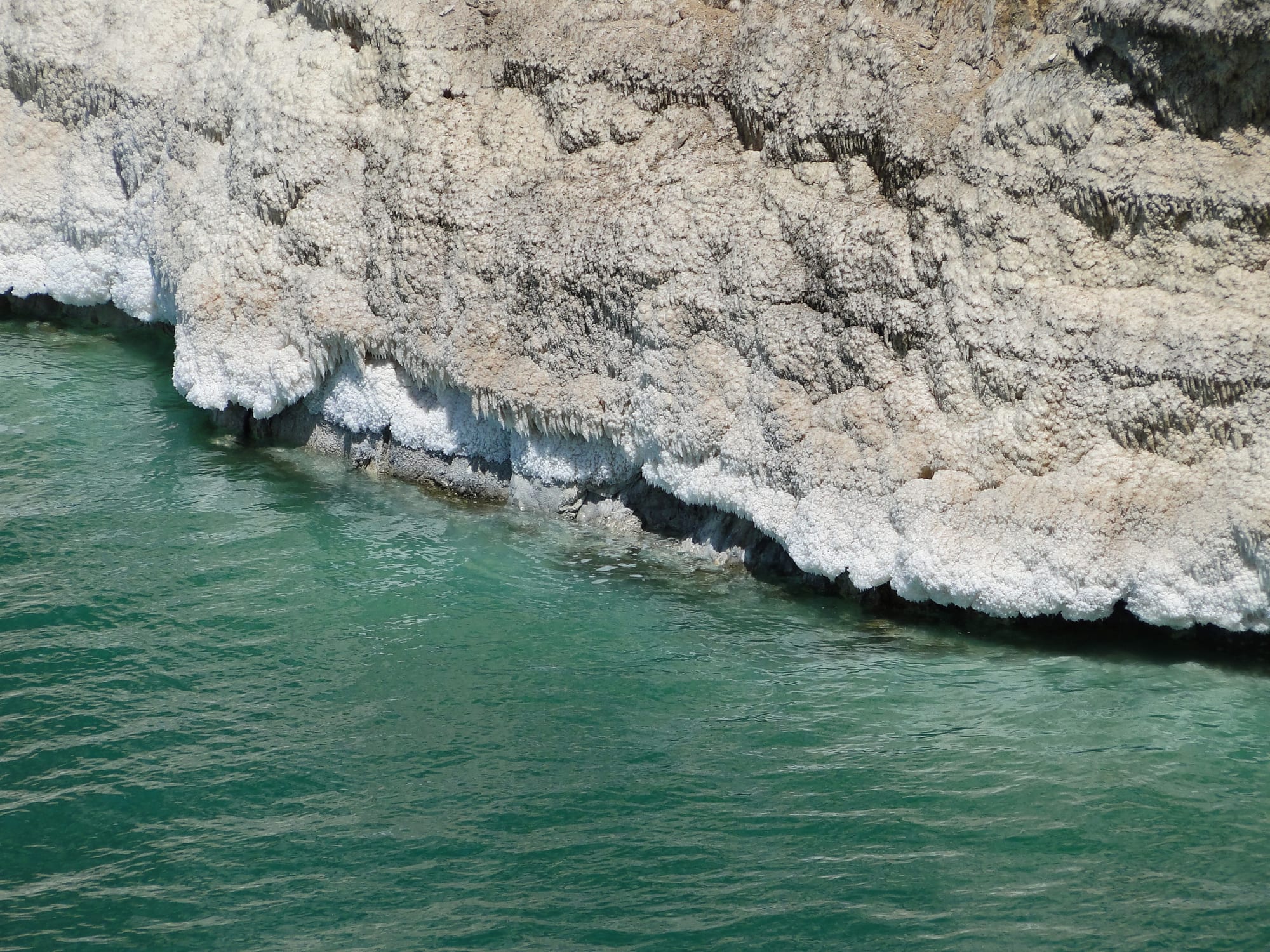 A calm blue sea meeting a salt-crusted shore with bare hills behind