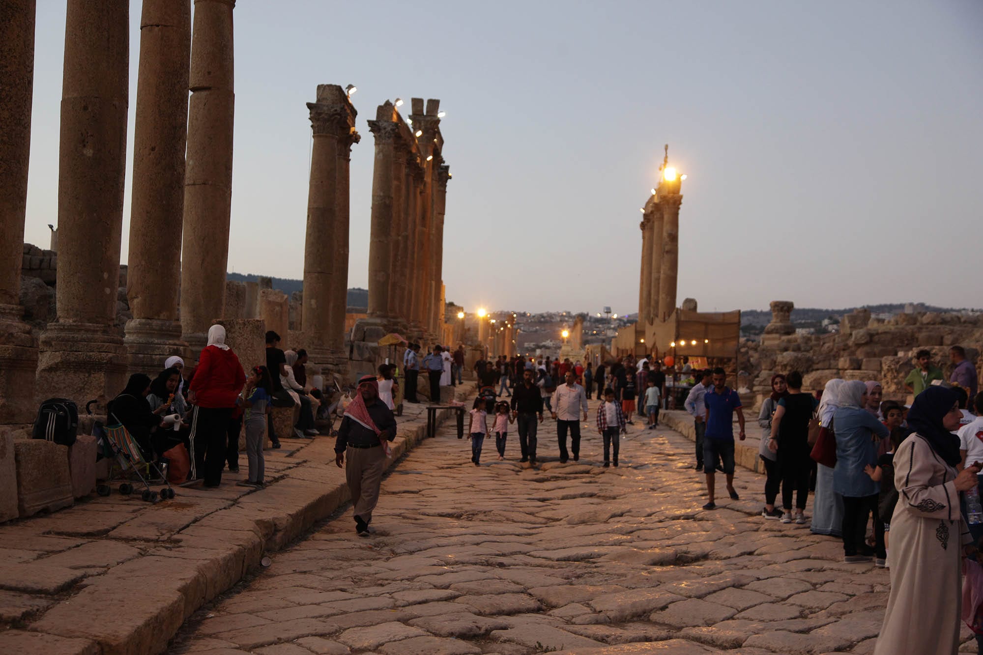 Families and visitors walk along an ancient paved street between tall Roman columns at dusk, with festival stalls and lights further along