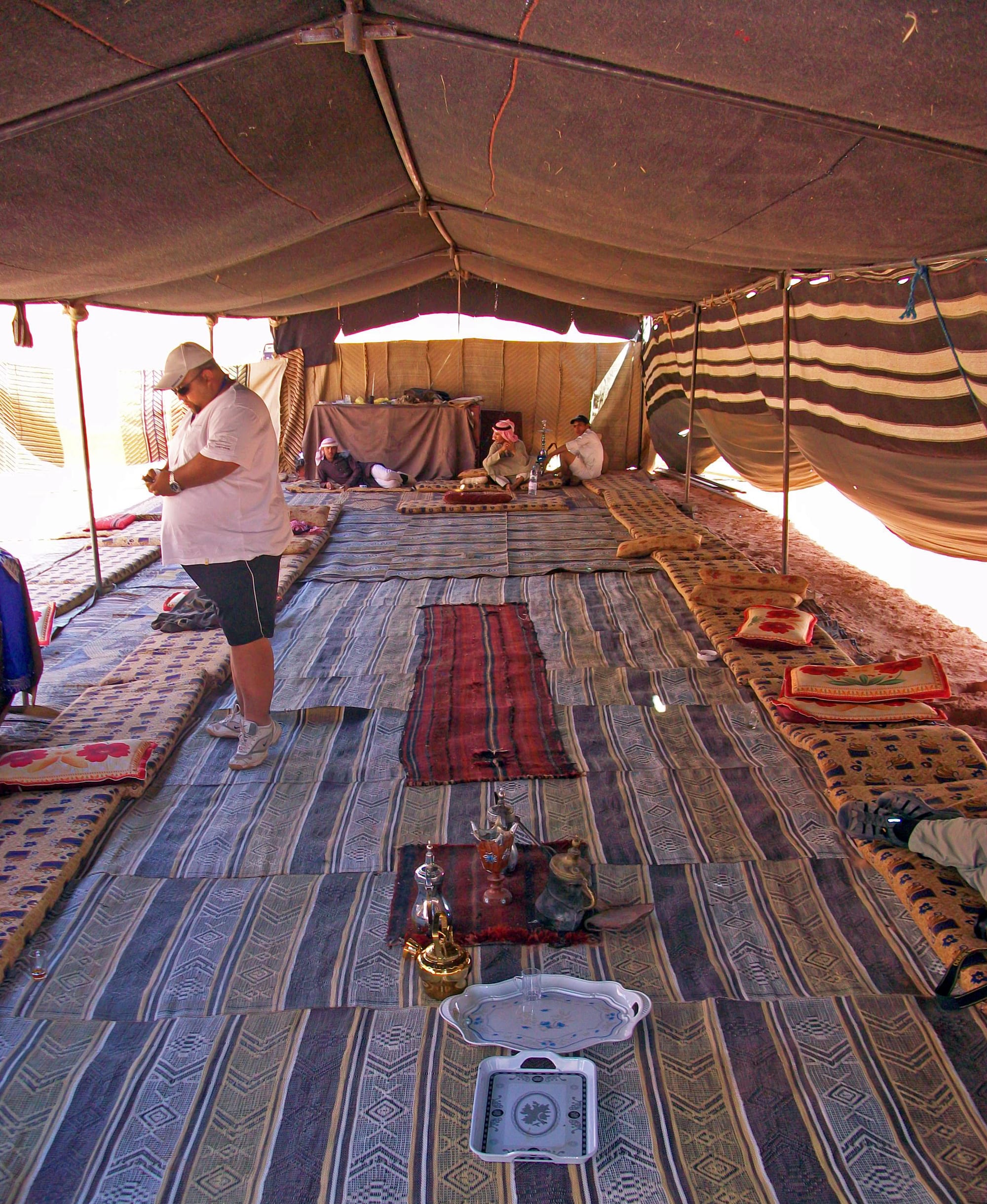 The inside of a long Bedouin tent laid with striped rugs and cushions along both sides, with coffee and tea pots and glasses set out on a cloth in the foreground