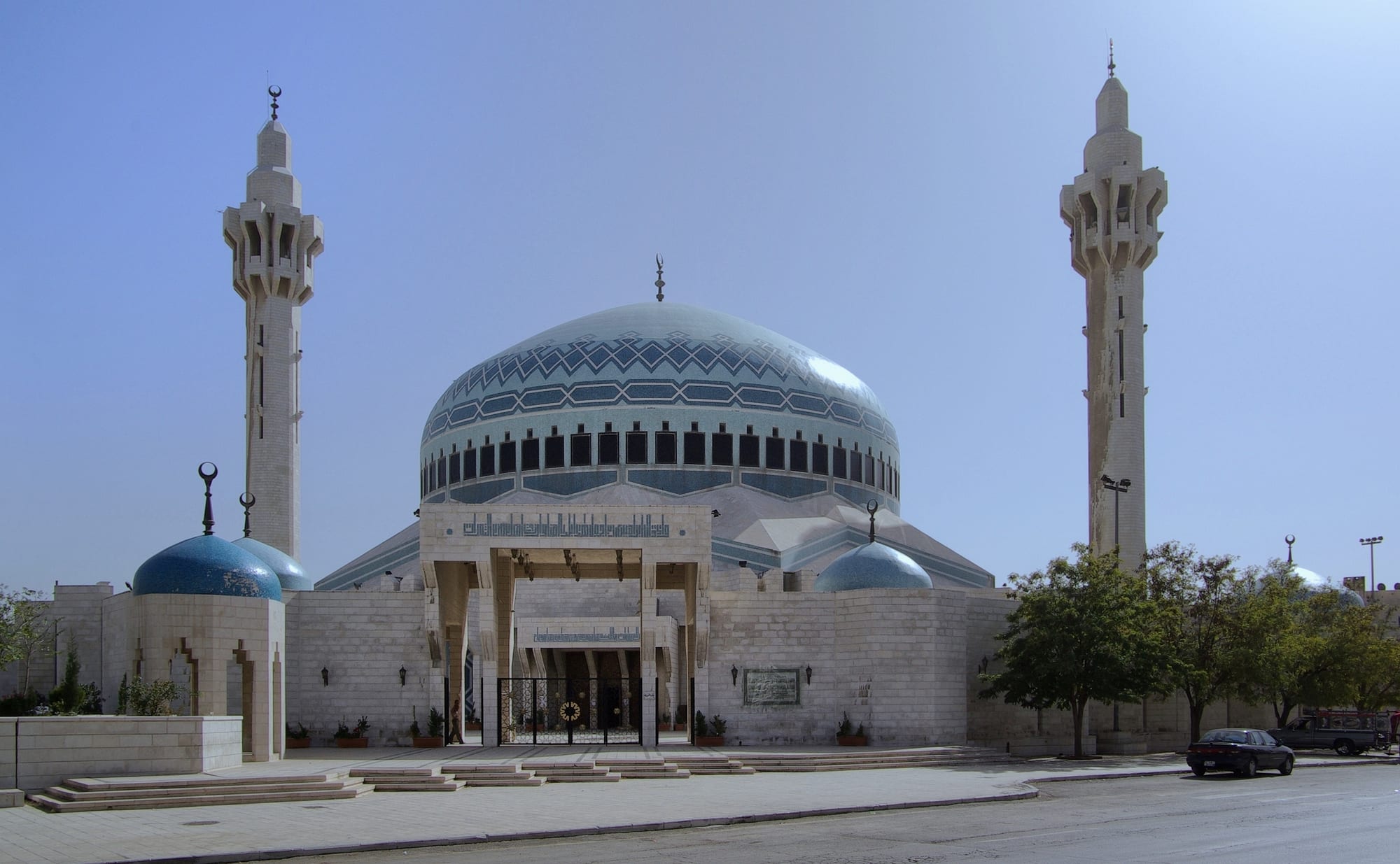 A white stone mosque with a large blue patterned dome ringed by small windows, two tall pale minarets and smaller blue domes, seen from a quiet street under a clear sky