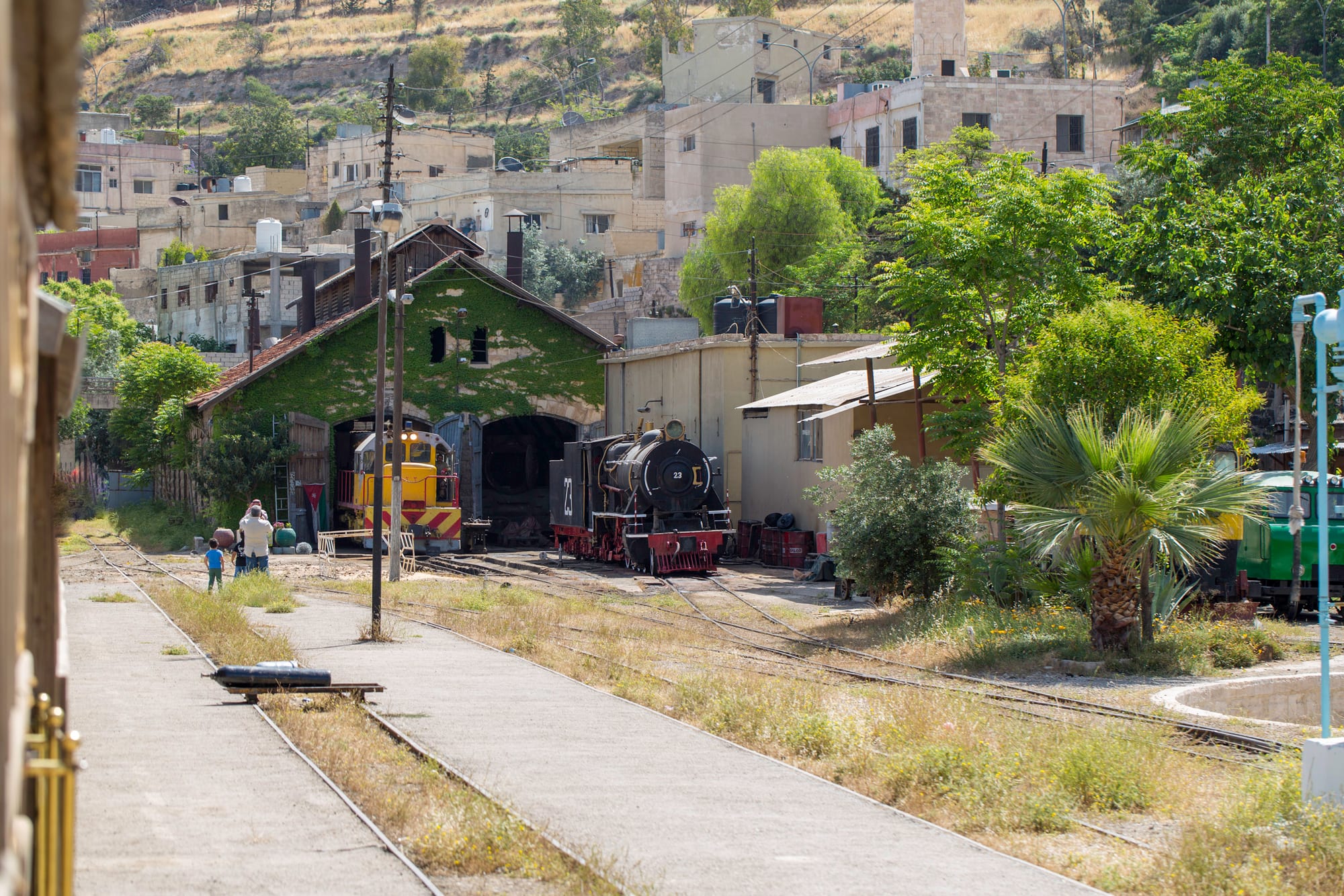 An old black steam locomotive and a yellow diesel engine outside a stone engine shed beside railway tracks, with houses on the hillside behind
