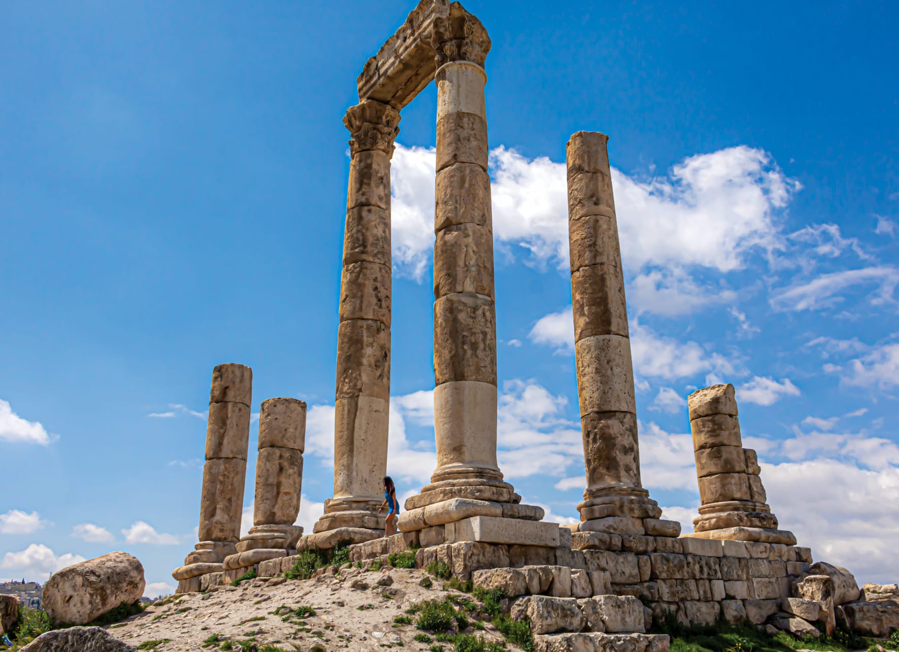 Tall weathered stone columns of a ruined ancient temple on a hilltop under a blue sky with white clouds
