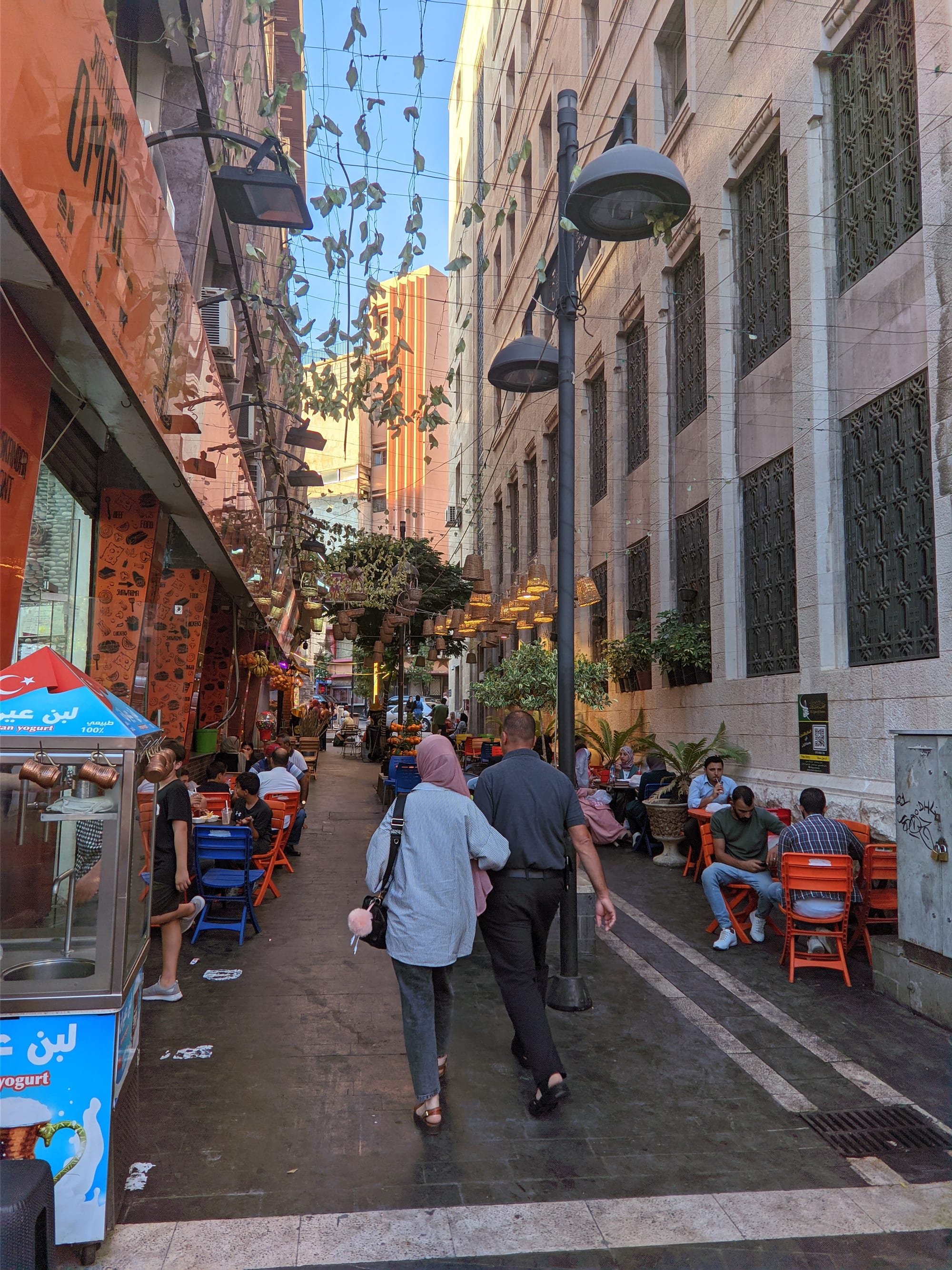 A pedestrian street in downtown Amman lined with cafes and orange chairs, with people walking past in early evening light
