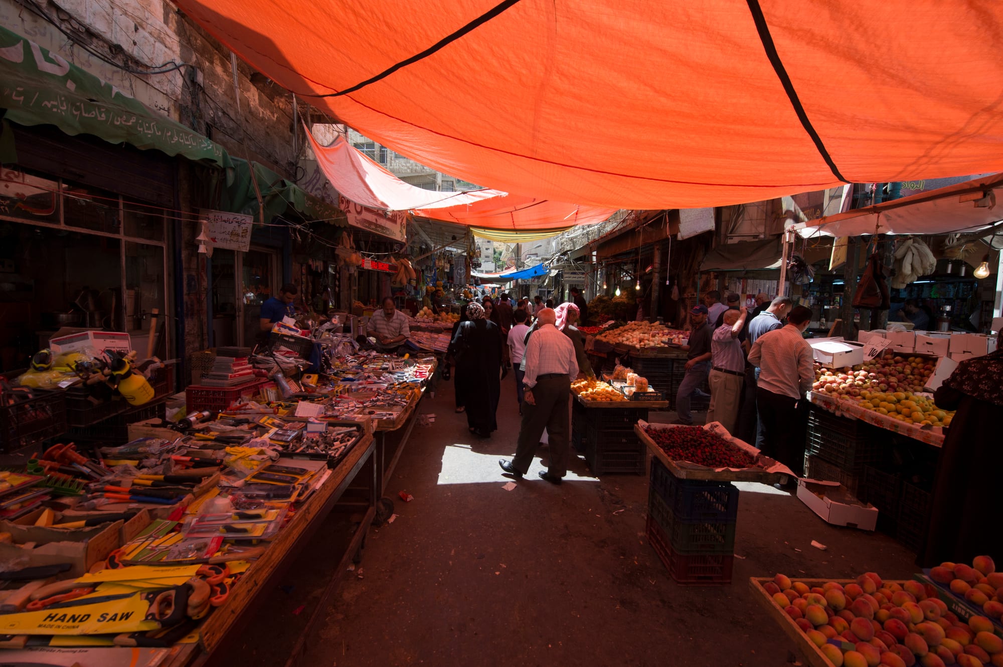 A covered market street in Amman with an orange awning overhead, shoppers browsing stalls of fruit and household goods