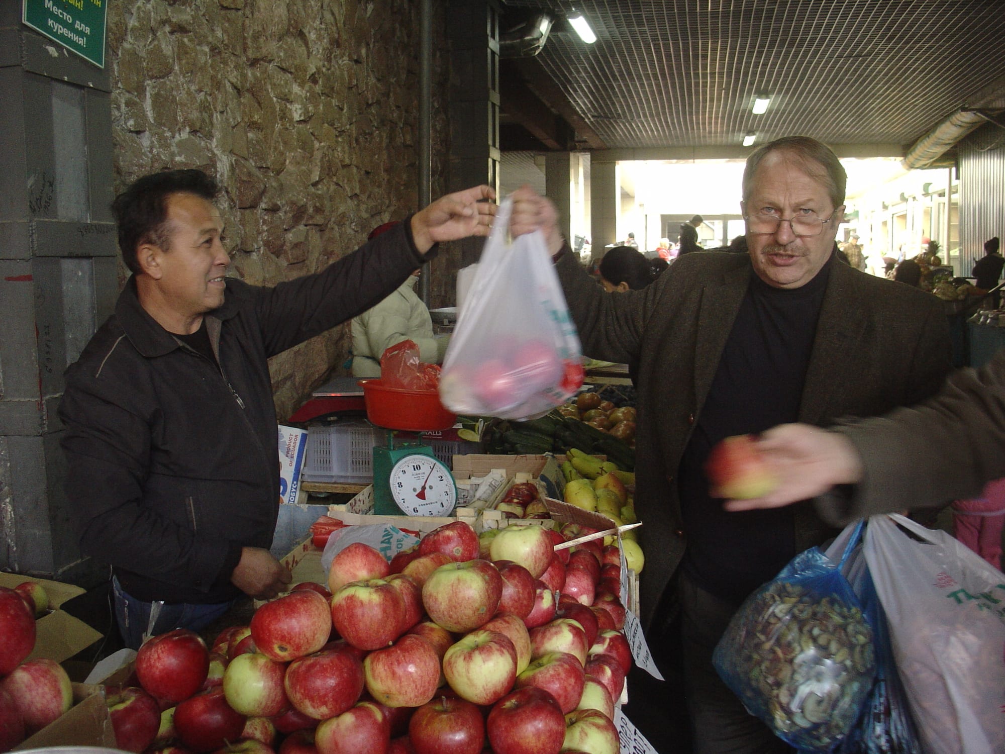 Two men at an indoor market stall, one handing the other a bag of red apples piled on the counter