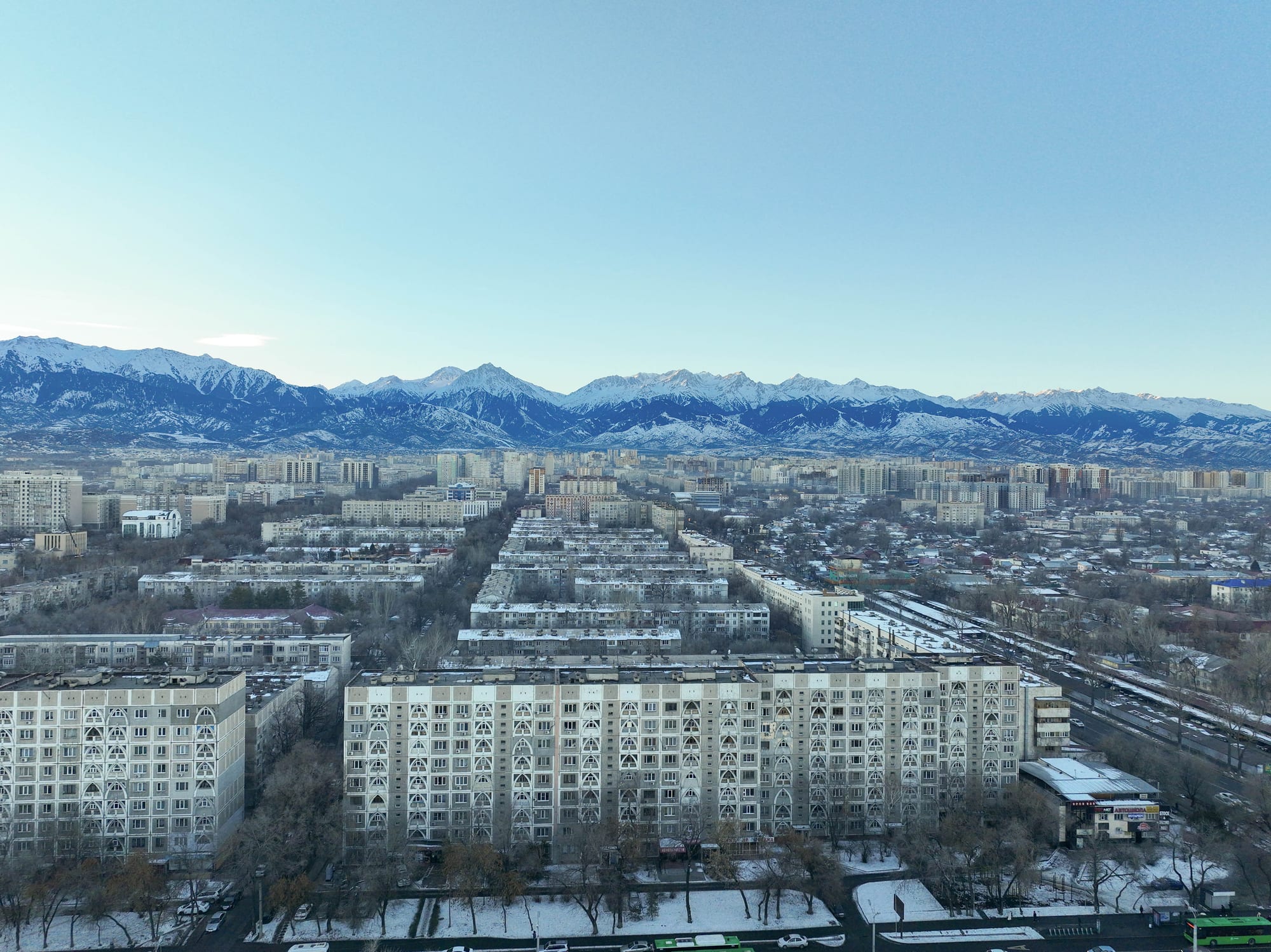 An aerial winter view of rows of Soviet-era apartment blocks in Almaty, with snow-capped Tian Shan mountains rising behind the city