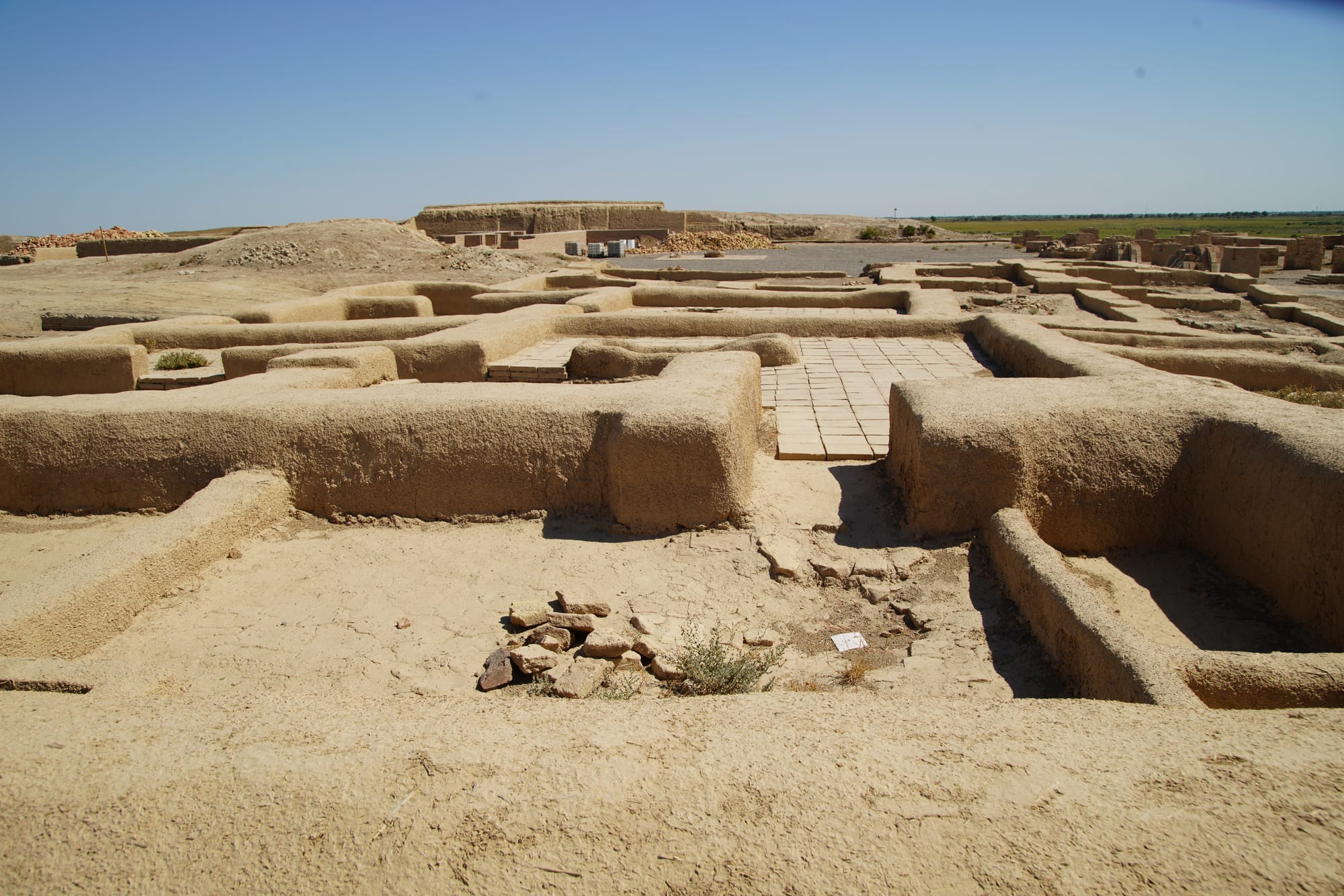 Low excavated mud-brick walls and a patch of paved floor at an archaeological site on a flat plain under a clear sky