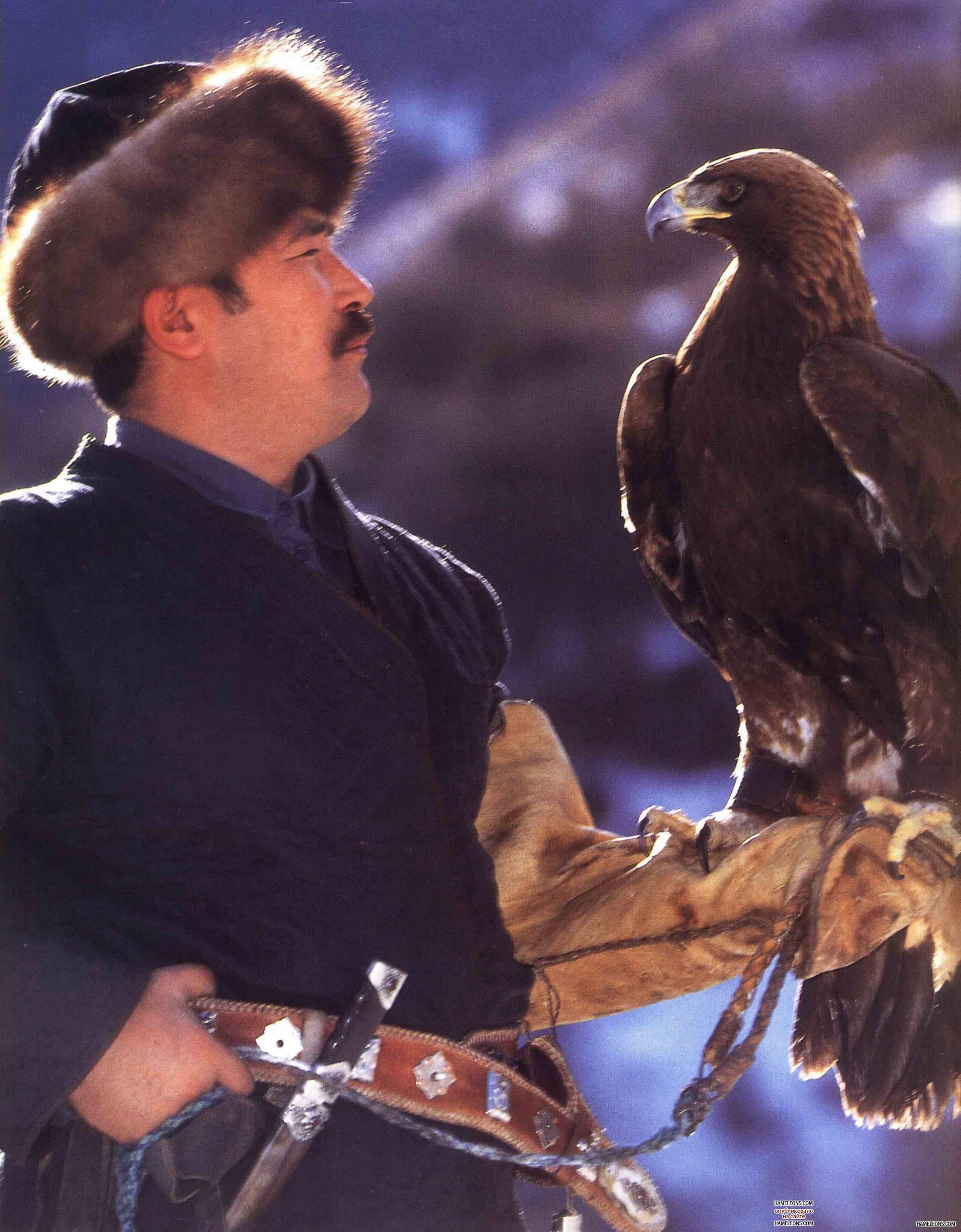 A Kazakh horseman in a fur hat and coat holding a large golden eagle on his gloved arm on the steppe