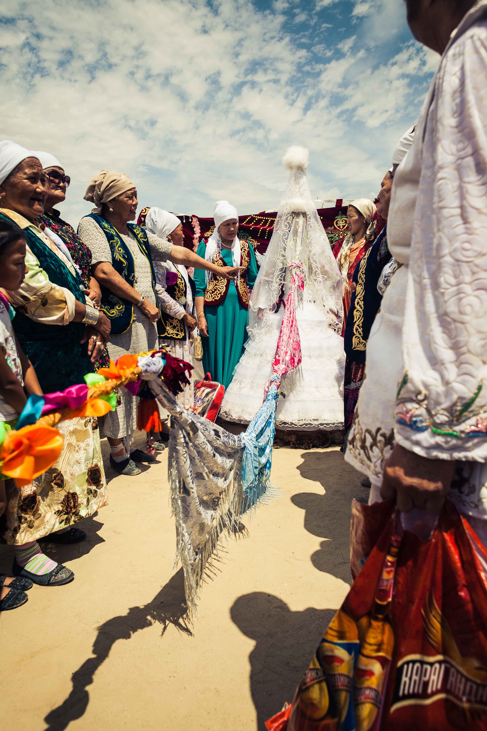 A bride in a white dress and tall white headdress with a long veil stands among older women in embroidered waistcoats and headscarves, under a bright sky