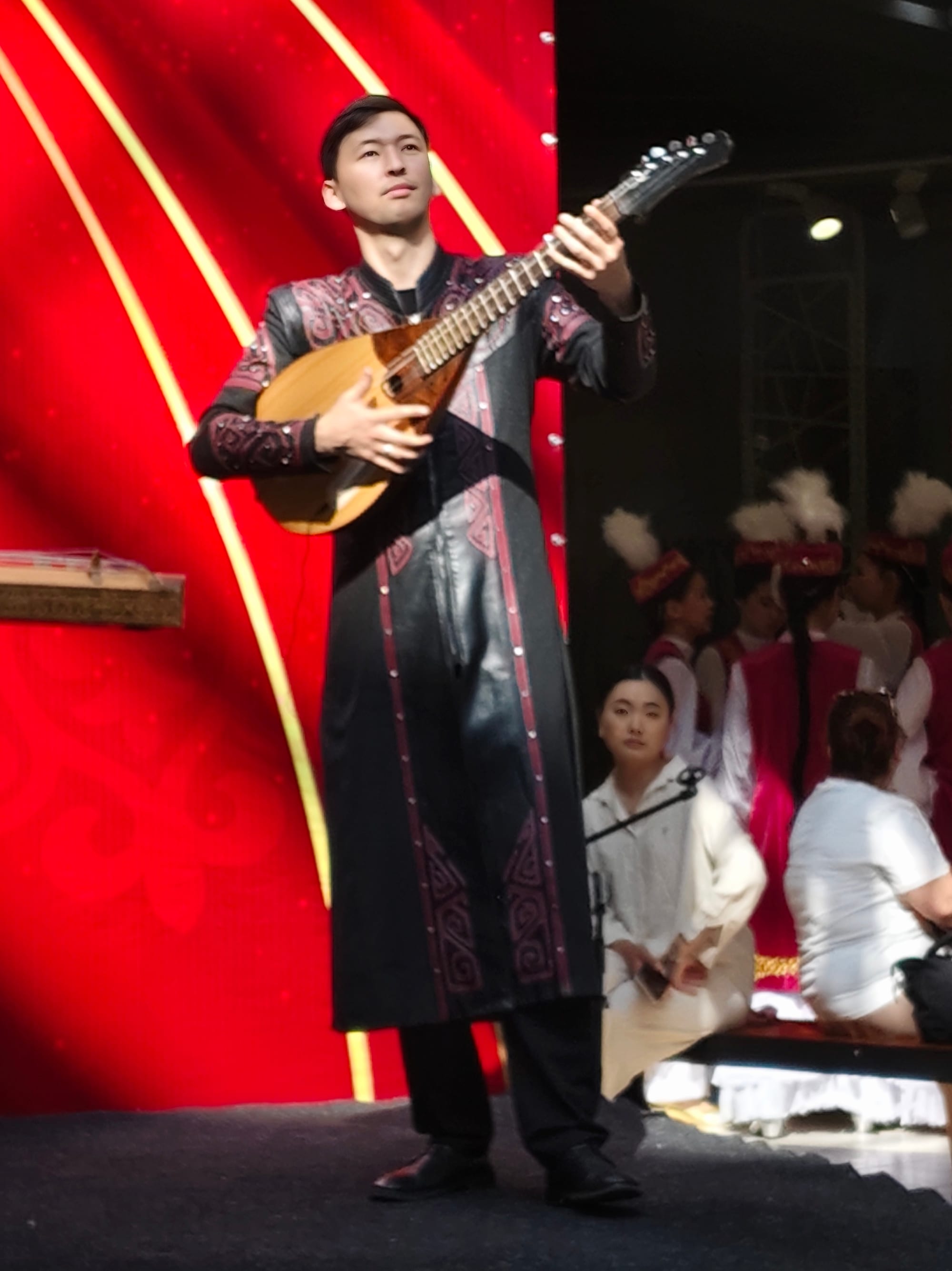 A musician in a long black coat embroidered with dark red patterns plays a stylised dombra on a stage with a red backdrop
