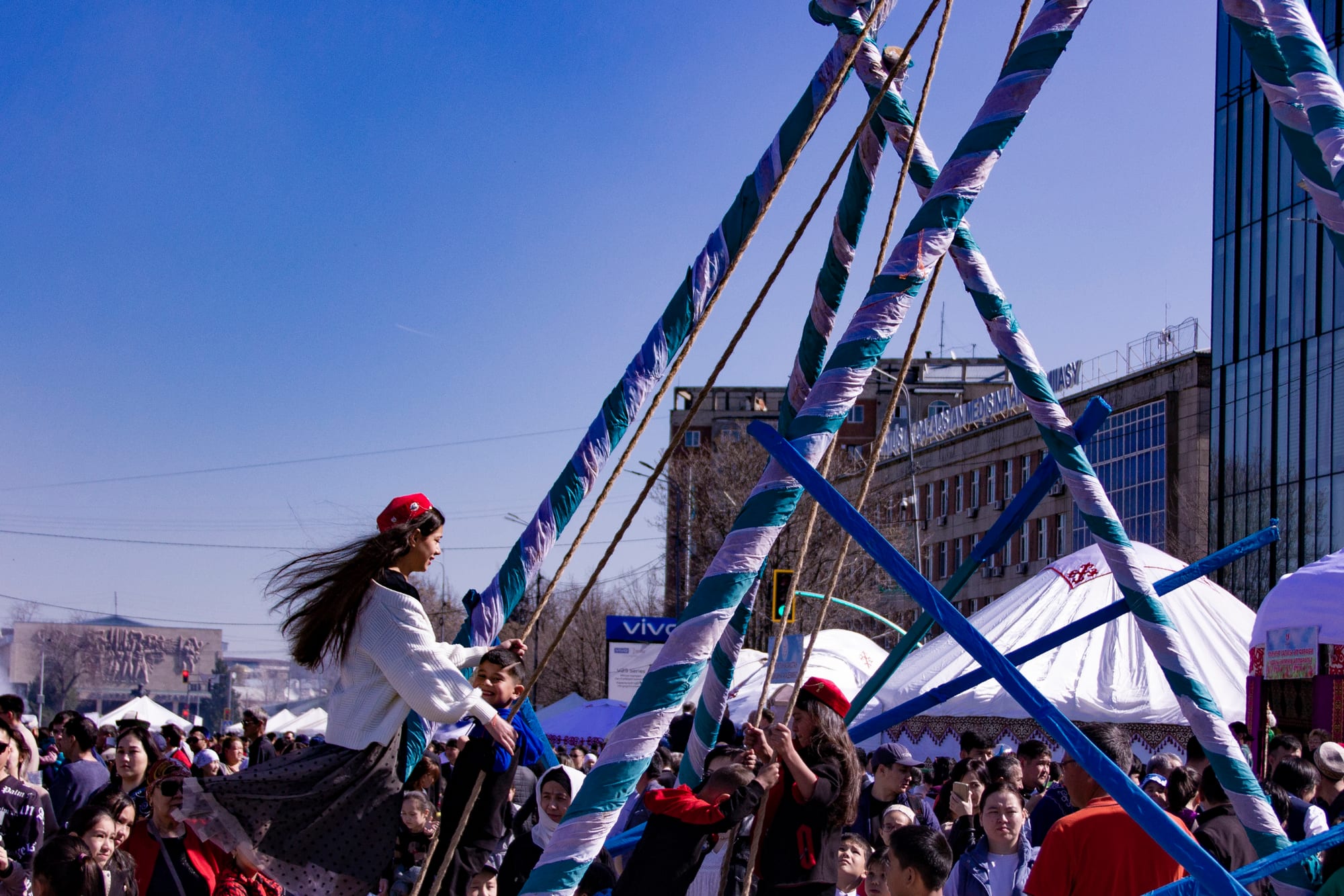 A young woman in a red cap and white jumper swings high on a tall frame of striped poles above a festival crowd, with white yurts and city buildings behind