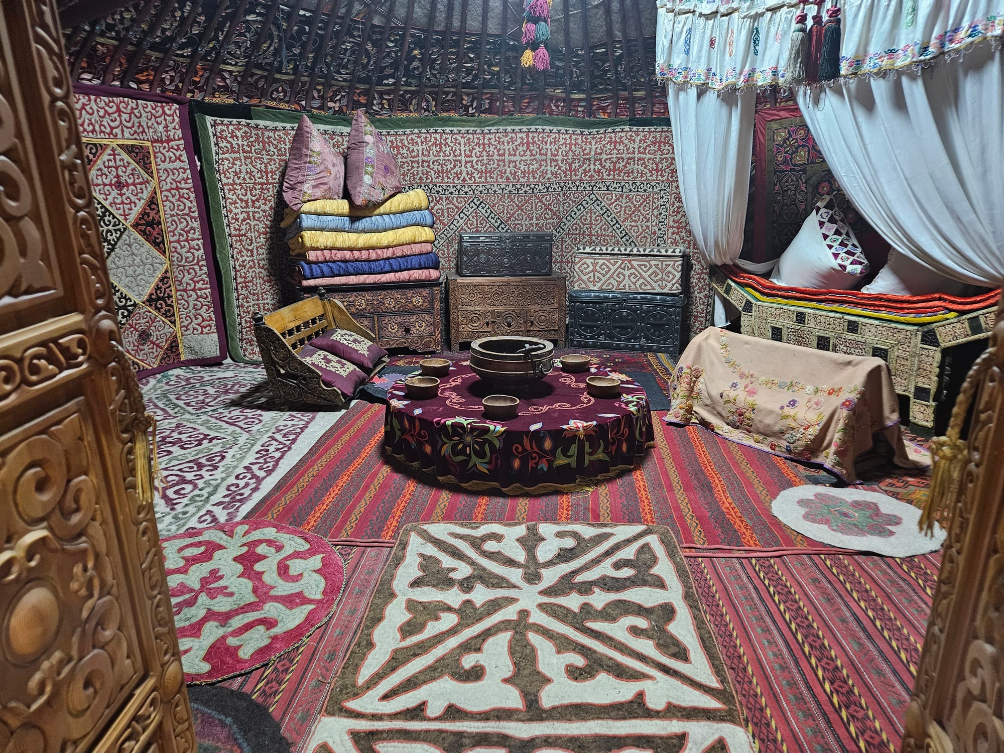 The inside of a yurt laid with red striped rugs and patterned felt carpets, with a low round table set with small bowls, carved chests and stacked quilts