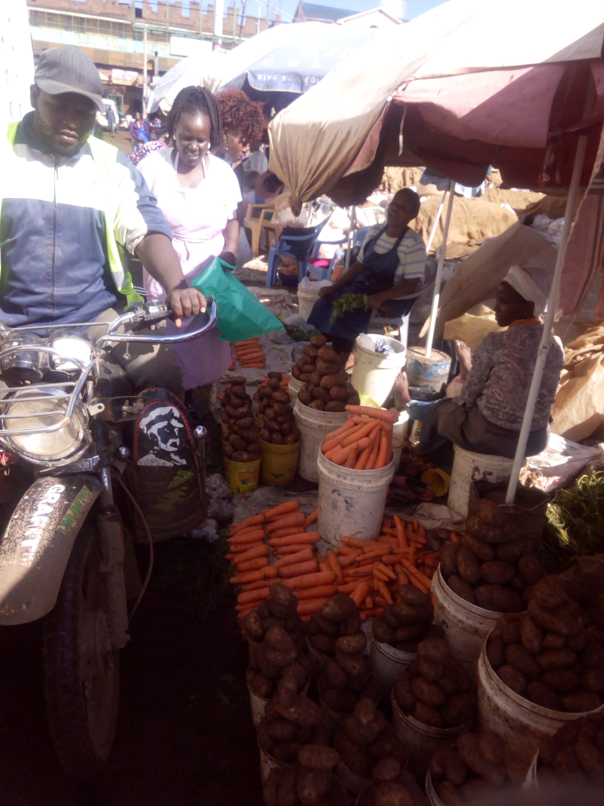 Vendors and customers crowded around buckets of carrots and potatoes at an open-air market stall under umbrellas