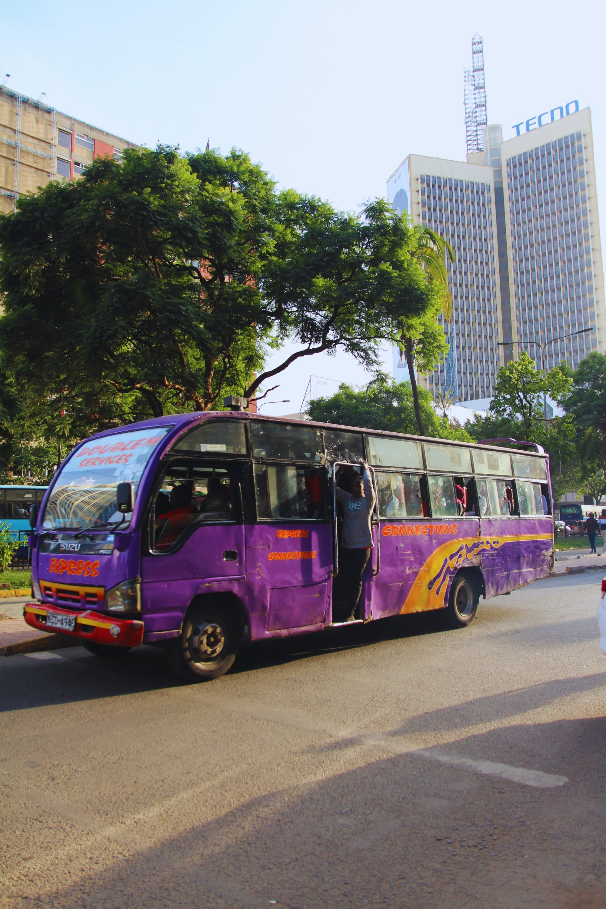 A purple and orange painted minibus with a passenger standing in its open doorway, stopped on a Nairobi street with high-rise towers behind