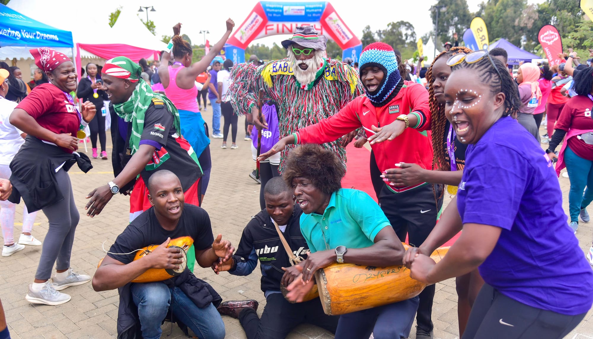 Three men crouch on paving beating long wooden drums while a crowd of smiling people in bright clothing, one in a shaggy costume and white beard, dance and point around them