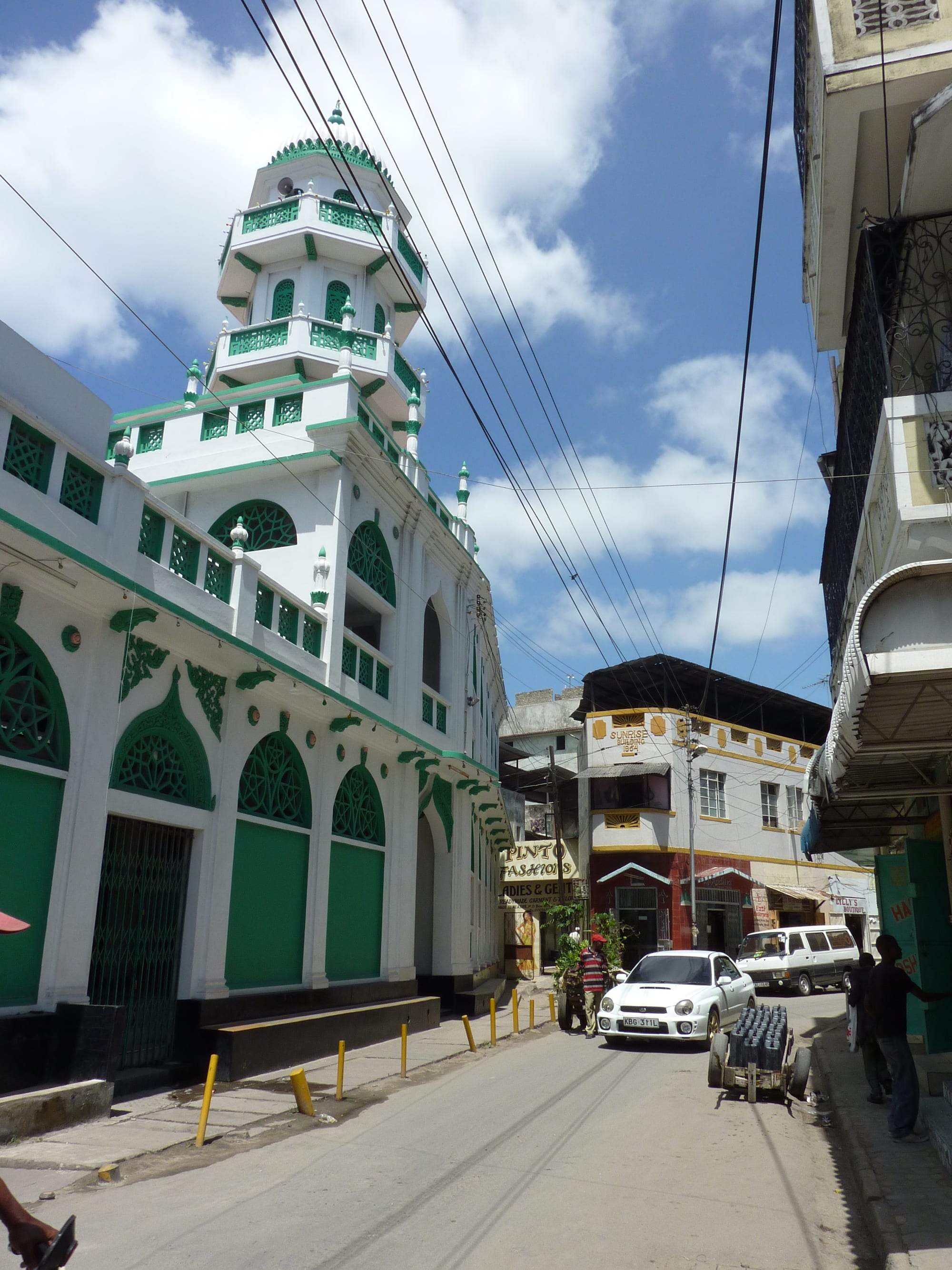 A white mosque with green latticework, green shutters and a tall tiered white and green minaret rises beside a street, with a white car and shopfronts beyond under a blue sky