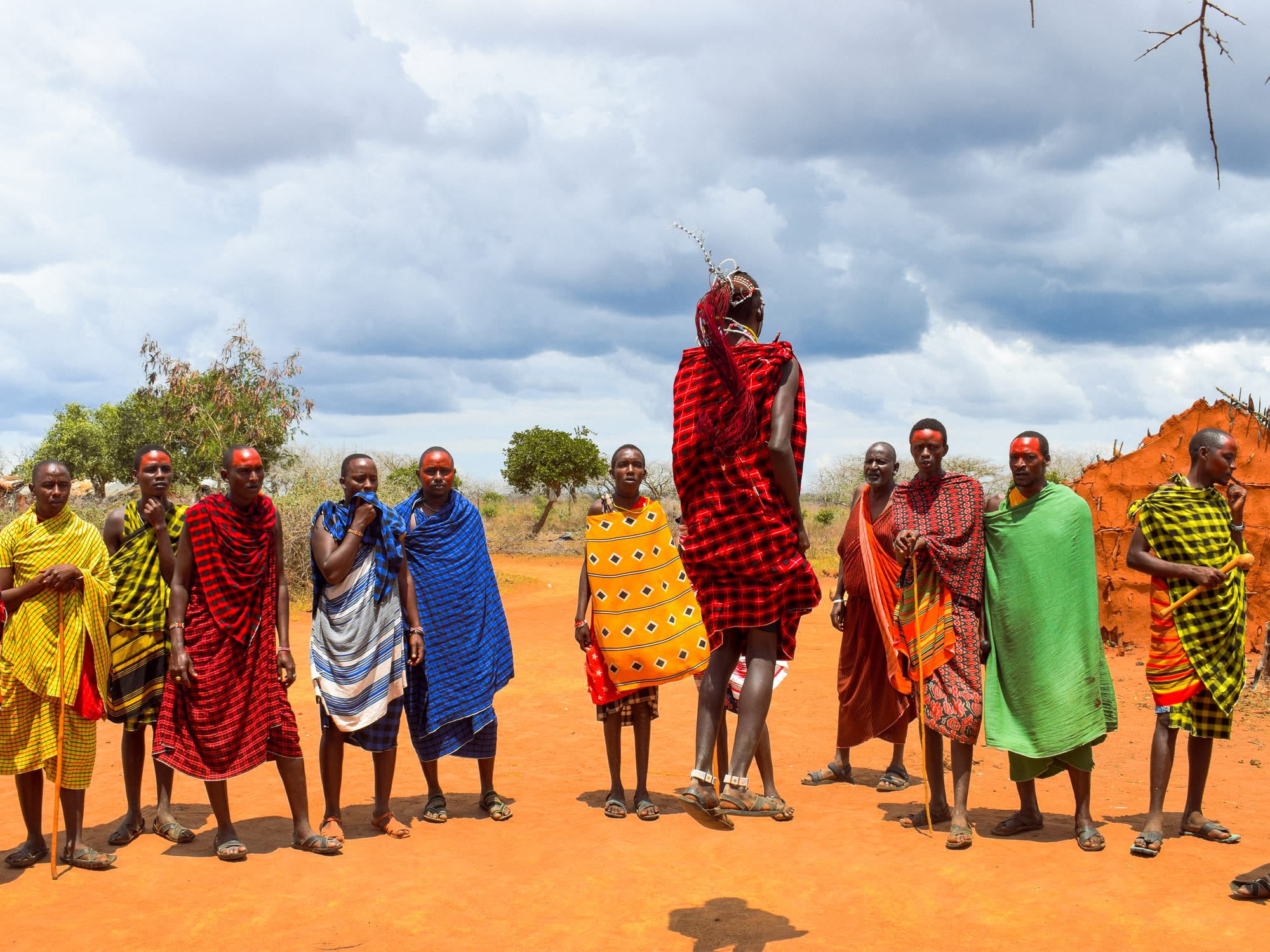 A line of Maasai men and women wrapped in bright red, yellow, blue and green checked and striped cloths stand on red earth under a cloudy sky as one man in a red checked cloth leaps high in the air