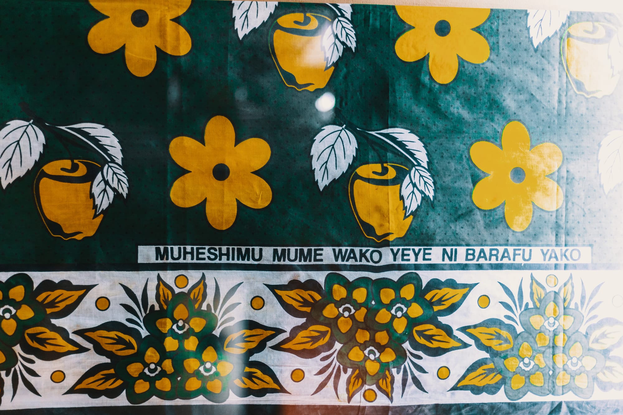 A green printed cloth with large yellow flowers and yellow apples with white leaves, above a white border of green and yellow flowers and a printed line of Swahili text
