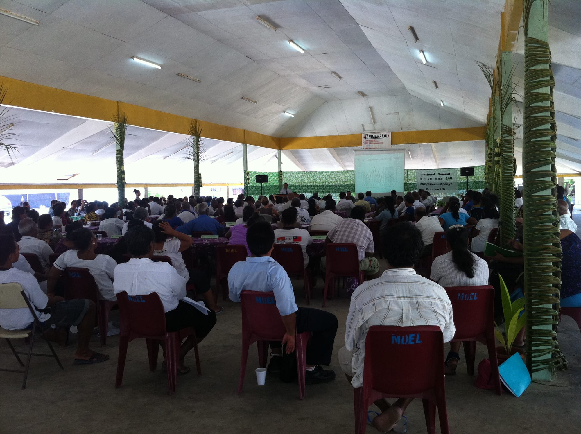 A large crowd seated on plastic chairs inside an open-sided thatched-roof maneaba meeting hall during a community gathering
