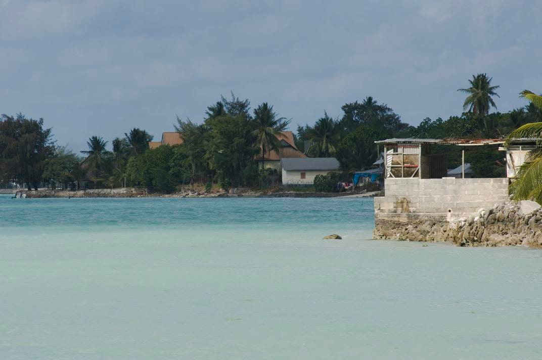 A lagoon-side village on South Tarawa with low houses and coconut palms along the shore, seen from the water at low tide