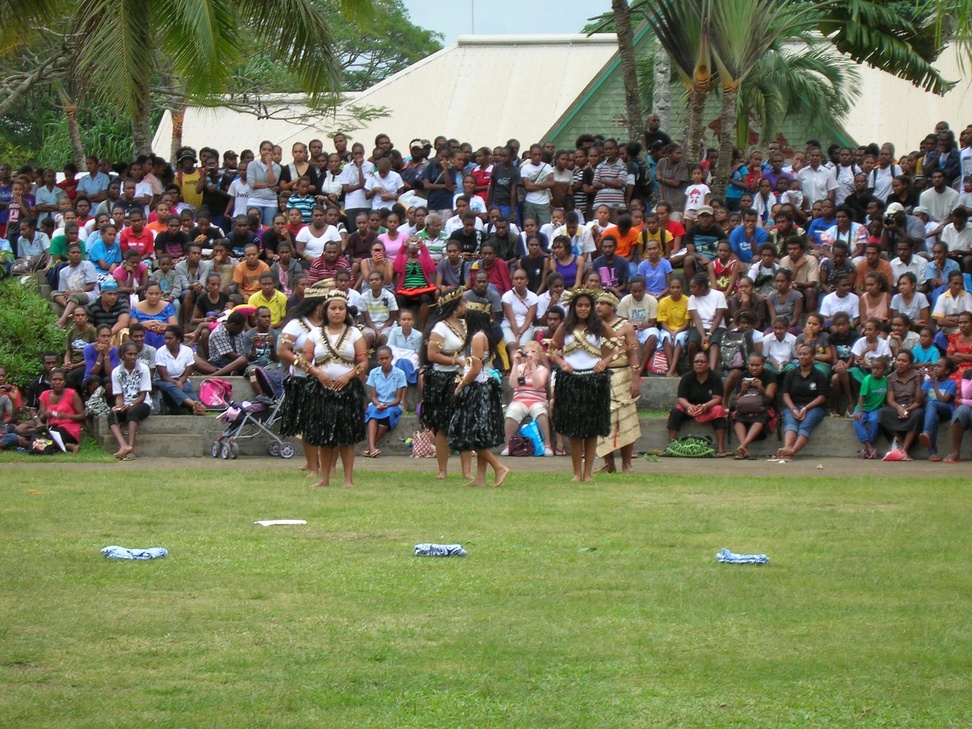 Four women dancers in white tops, black grass skirts and woven headbands perform on a lawn beside a man in a woven skirt, watched by a large crowd seated on steps